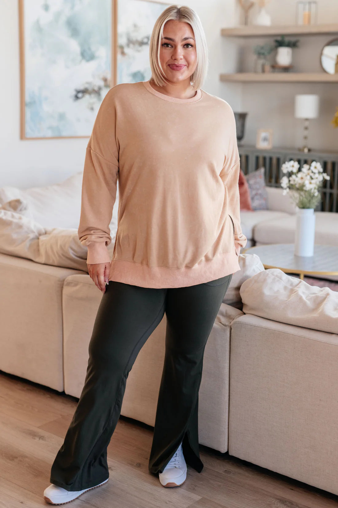 Smiling woman with straight blonde hair wearing a relaxed fit khaki sweatshirt paired with black pants in a bright living room setting.
