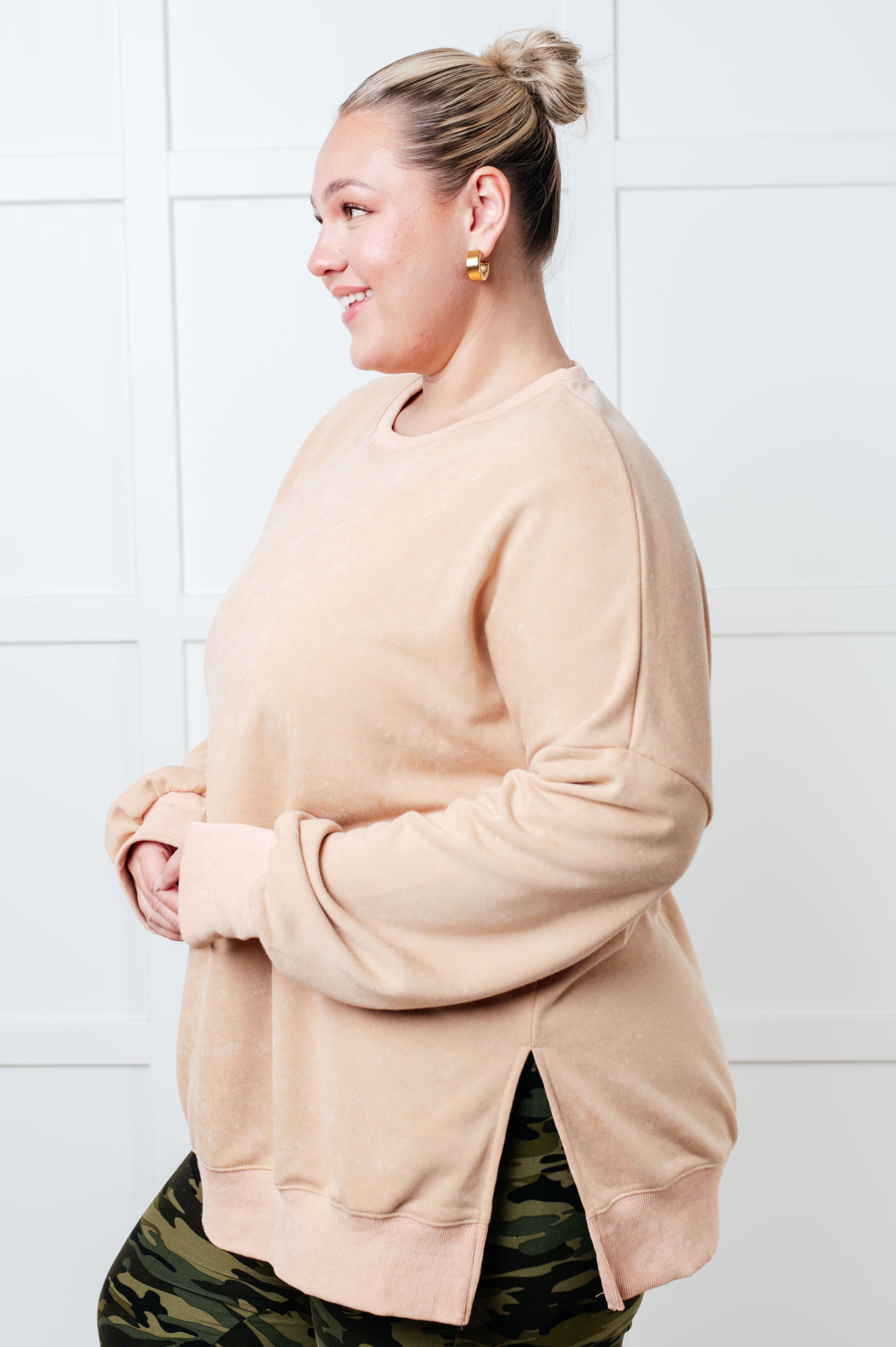 Smiling woman wearing a relaxed khaki oversized sweatshirt with side slits paired with camo leggings, standing against a white paneled wall.