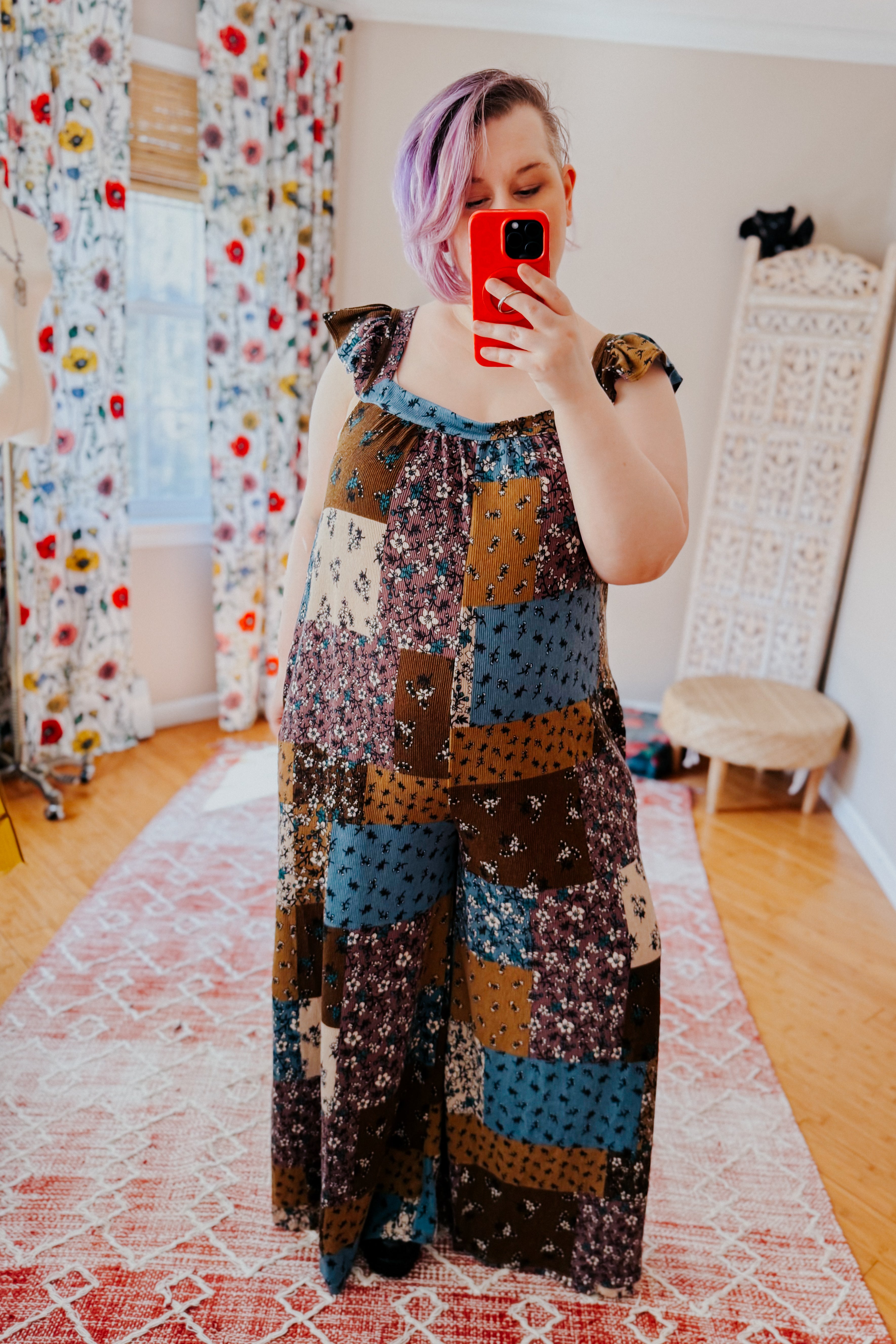 Woman with short lavender hair taking a mirror selfie wearing a loose-fitting patchwork jumpsuit with ruffle straps in brown, blue, and cream tones.