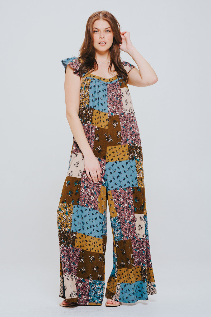 Woman with long brown hair wearing a relaxed fit patchwork jumpsuit with ruffle sleeves in brown, blue, and mustard tones against a light background.