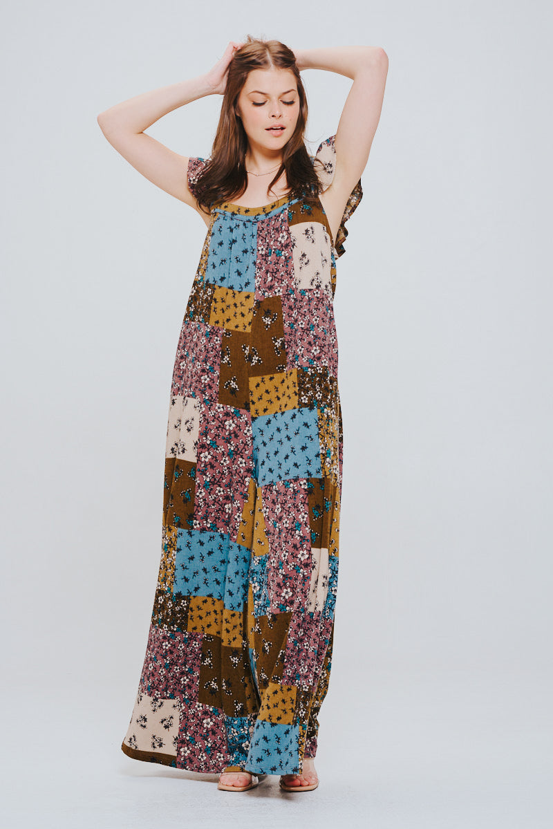 Woman with long brown hair wearing a sleeveless patchwork jumpsuit in brown, blue, mustard, and cream tones with ruffle details, standing with eyes closed and hands raised behind her head.