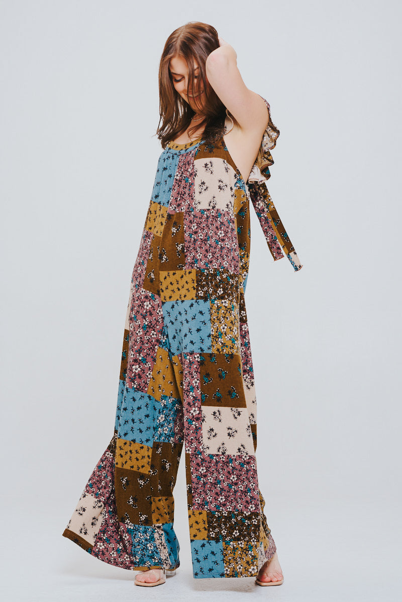 Woman wearing a loose-fitting patchwork jumpsuit in brown, blue, mustard, and cream tones with ruffle details on the shoulders, standing with one arm raised.