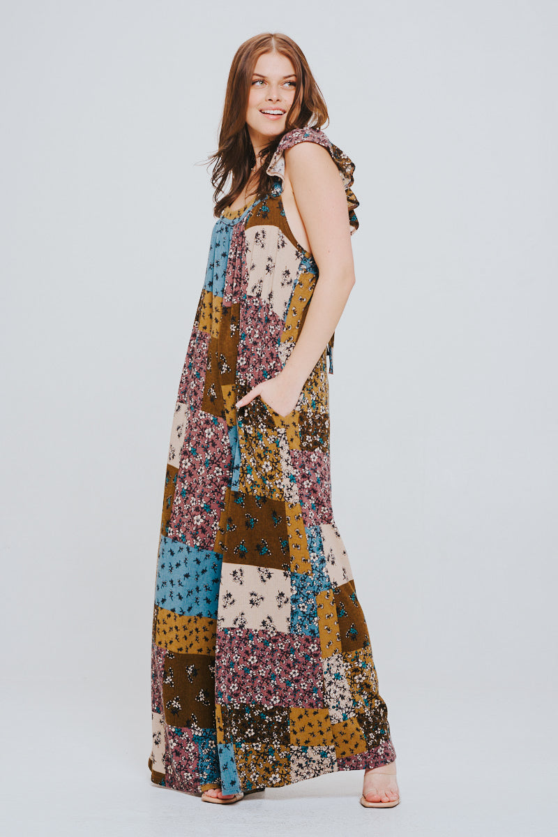Young woman wearing a sleeveless knit patchwork jumpsuit in brown, blue, and cream tones with ruffle details on the shoulders, standing with hands in pockets and smiling.
