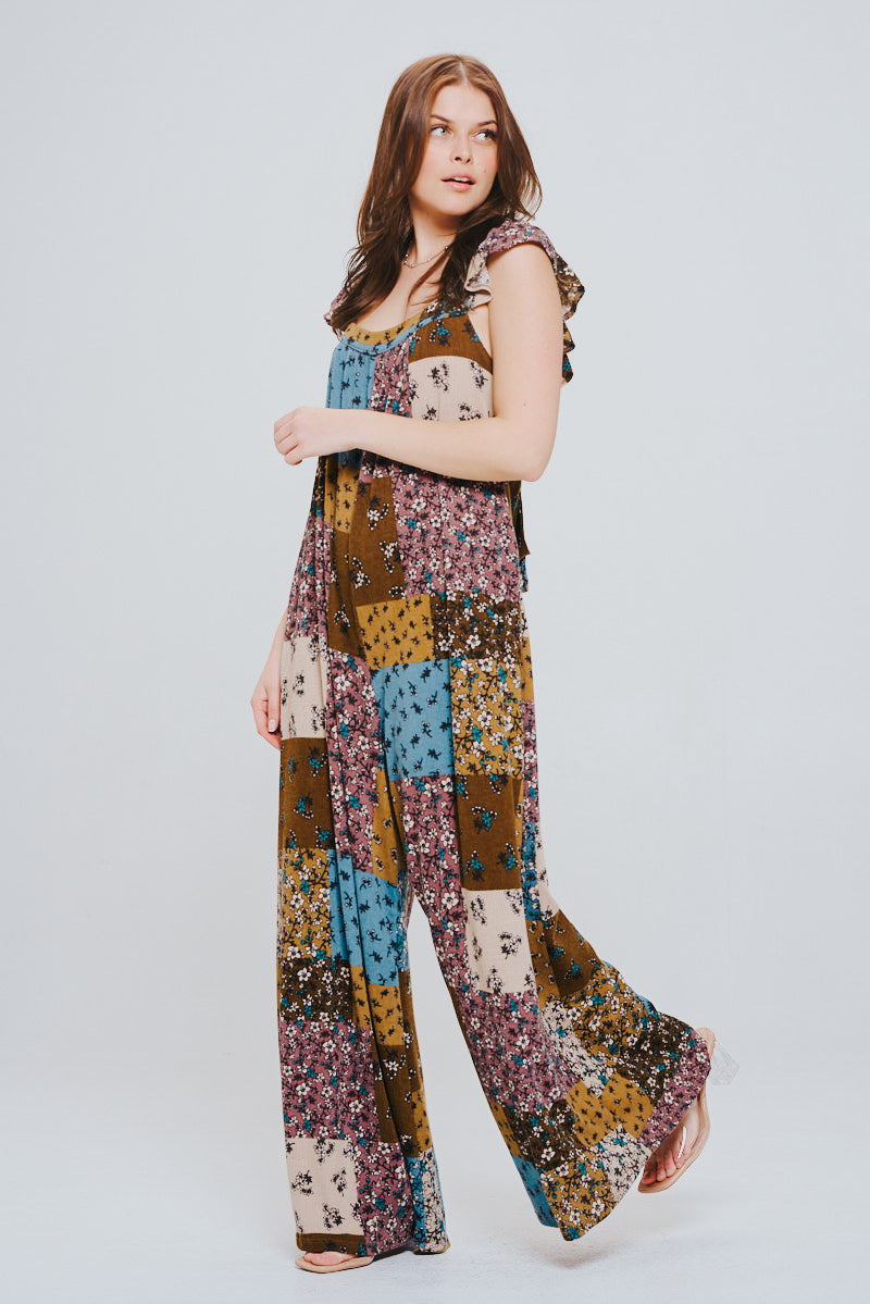 Woman with long brown hair wearing a flowy knit patchwork jumpsuit in various brown, blue, and cream tones with ruffle shoulder details, standing and looking to the side.