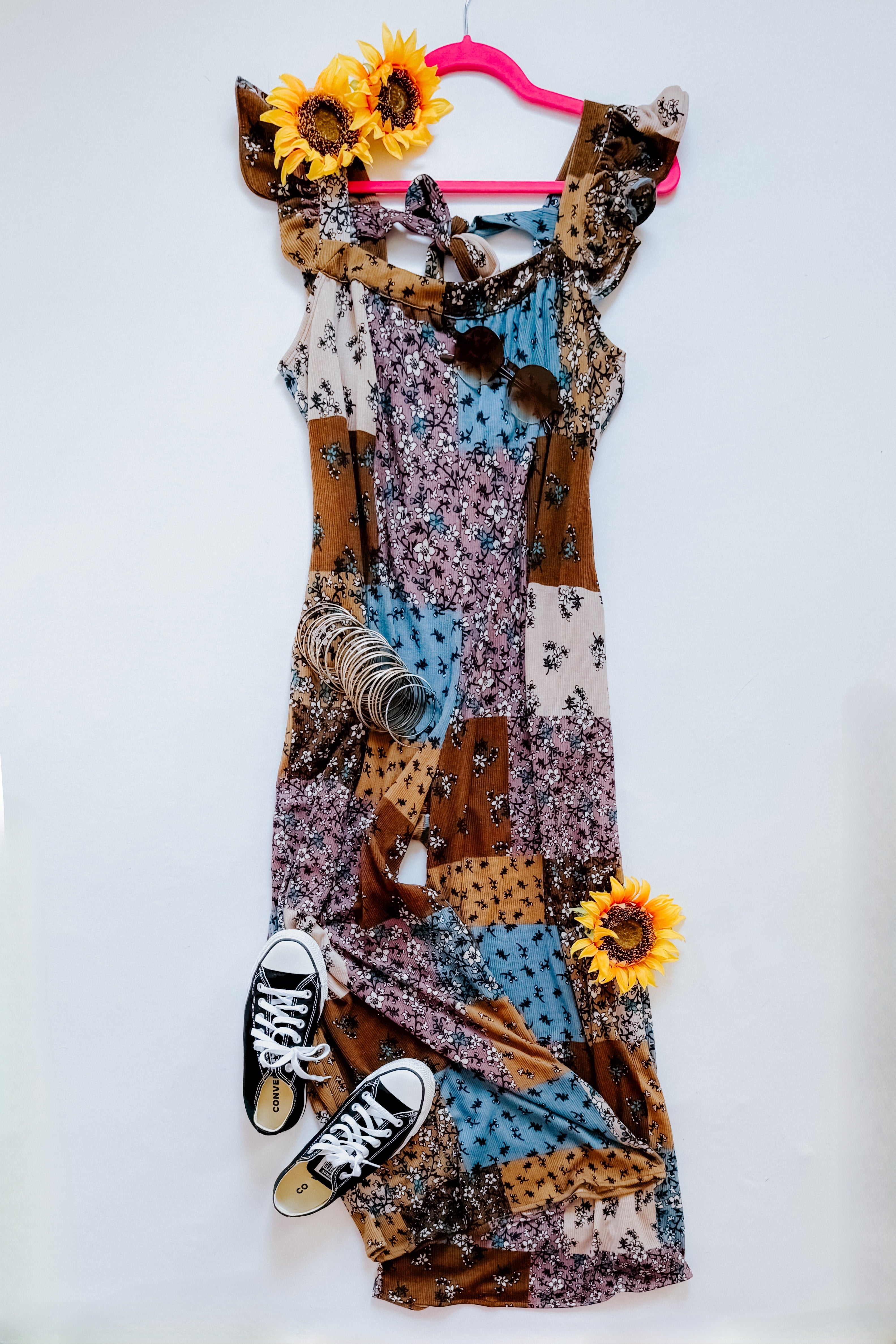 Sleeveless knit patchwork dress in brown, blue, and cream tones with ruffle shoulder details, styled with black sneakers and sunflowers.