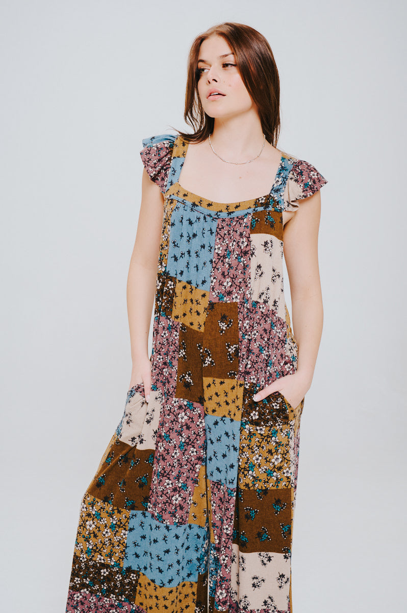 Woman with long brown hair wearing a sleeveless knit jumpsuit in brown, blue, and mustard patchwork with ruffle shoulder details and pockets.