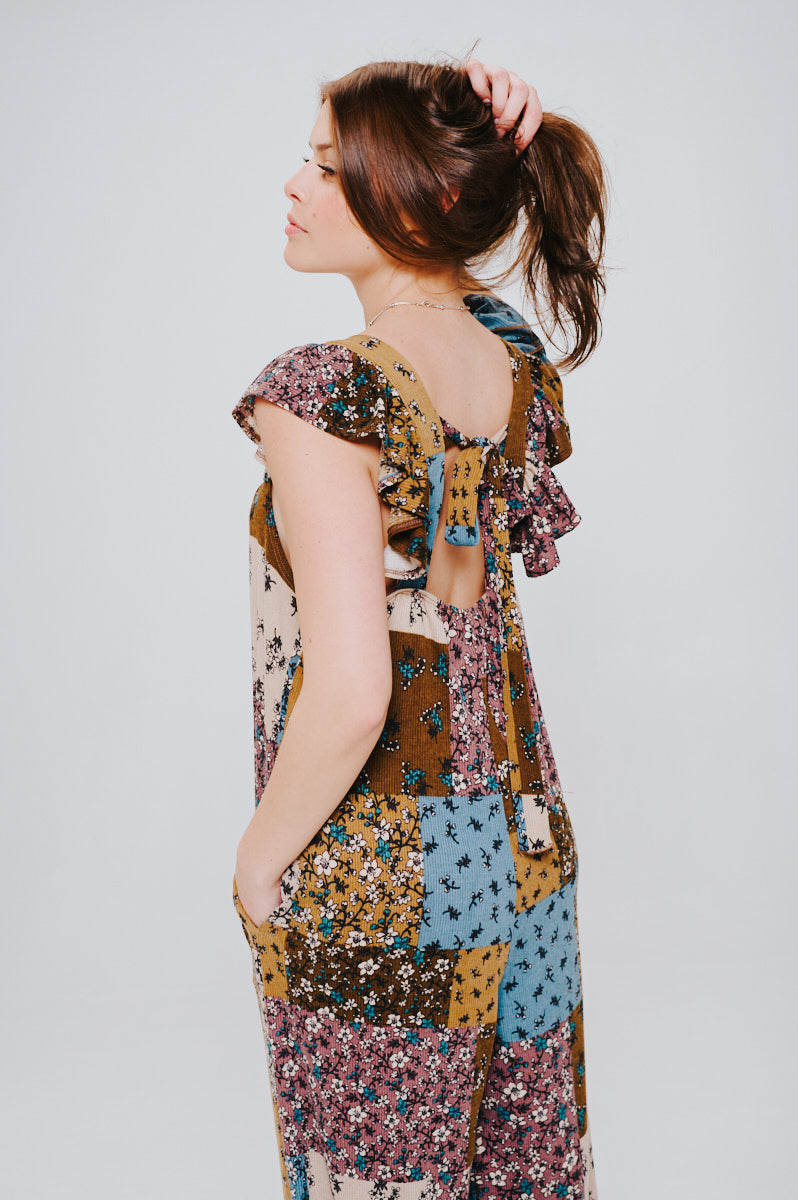 Woman with brown hair styled in a ponytail wearing a sleeveless knit patchwork jumpsuit in brown, blue, and cream tones with ruffled shoulder details and a bow tied at the back.