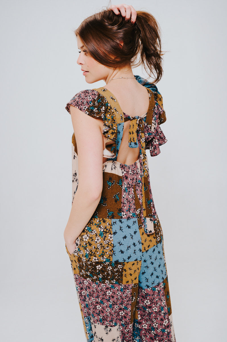 Woman with brown hair in a loose patchwork jumpsuit featuring ruffle sleeves, a tie-back detail, and a mix of brown, blue, and floral patterns.