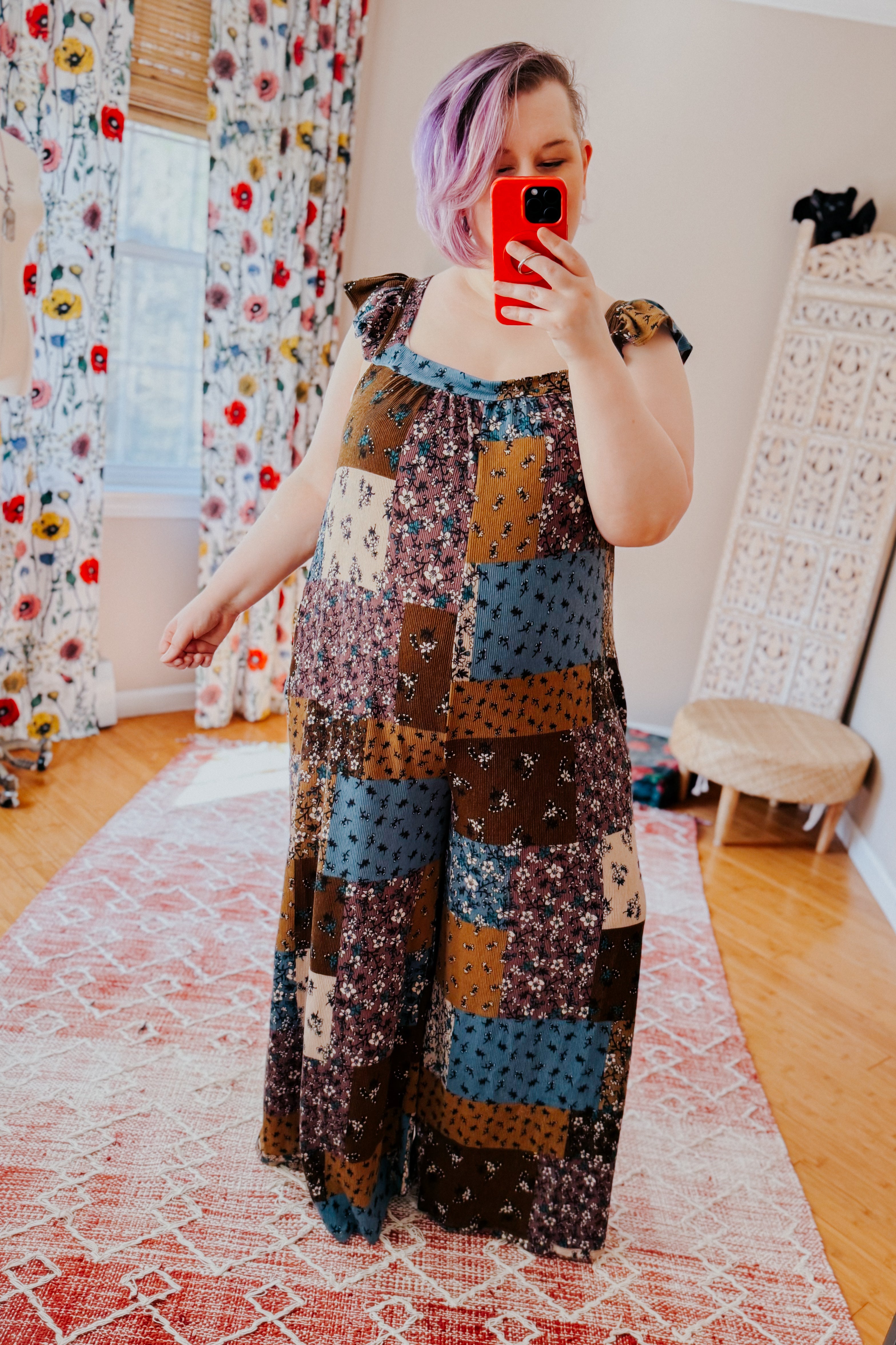 Woman with short lavender hair taking a mirror selfie wearing a loose, flowy patchwork jumpsuit with ruffle tie straps in brown, cream, and dusty blue tones.