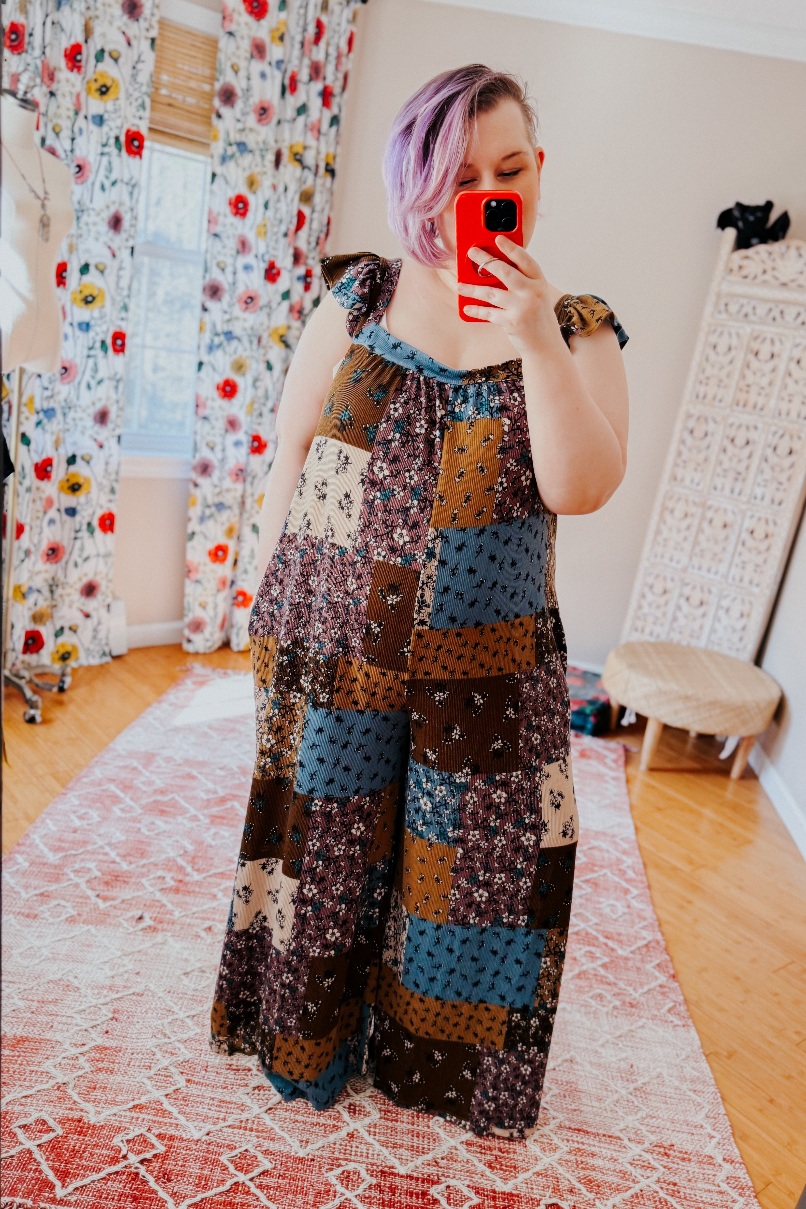 Woman taking a mirror selfie wearing a sleeveless patchwork jumpsuit with ruffle shoulder ties in brown, blue, and cream knit fabric.