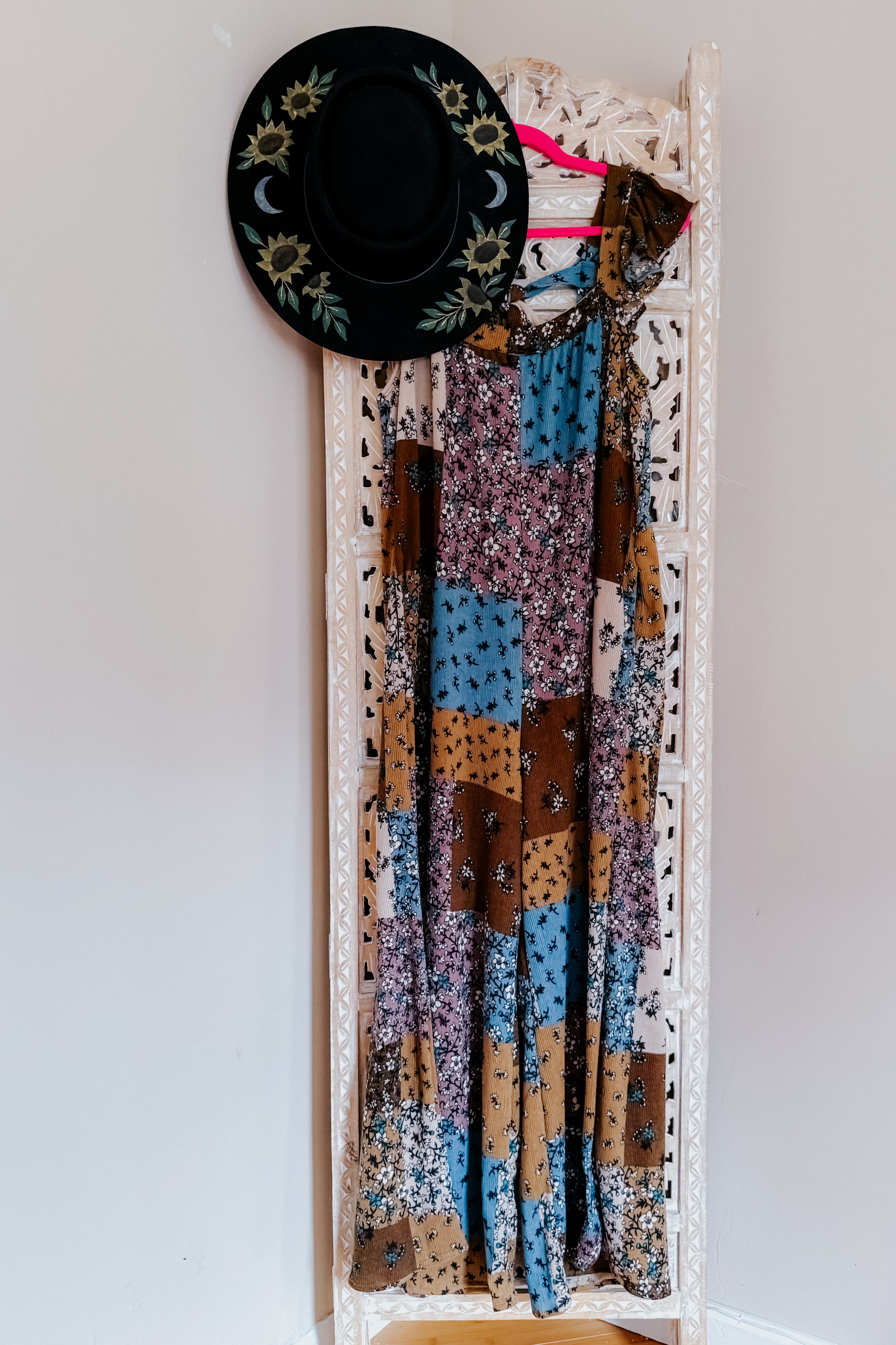 Sleeveless knit jumpsuit with a mix of floral and patchwork patterns in brown, blue, and cream tones hanging on a decorative white screen alongside a black hat with sunflower embroidery.