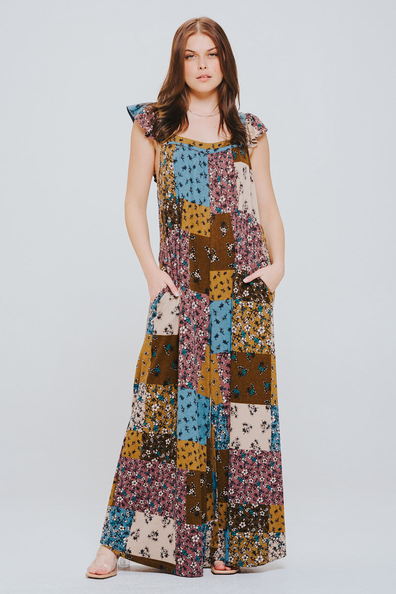 Woman with long brown hair wearing a colorful patchwork knit jumpsuit with ruffle sleeves, standing with hands in pockets against a plain background.