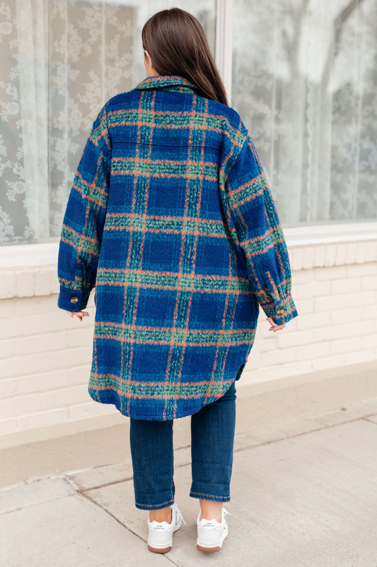 Back view of a woman wearing a blue plaid brushed flannel coat with a scooped hem over jeans and white sneakers by a window