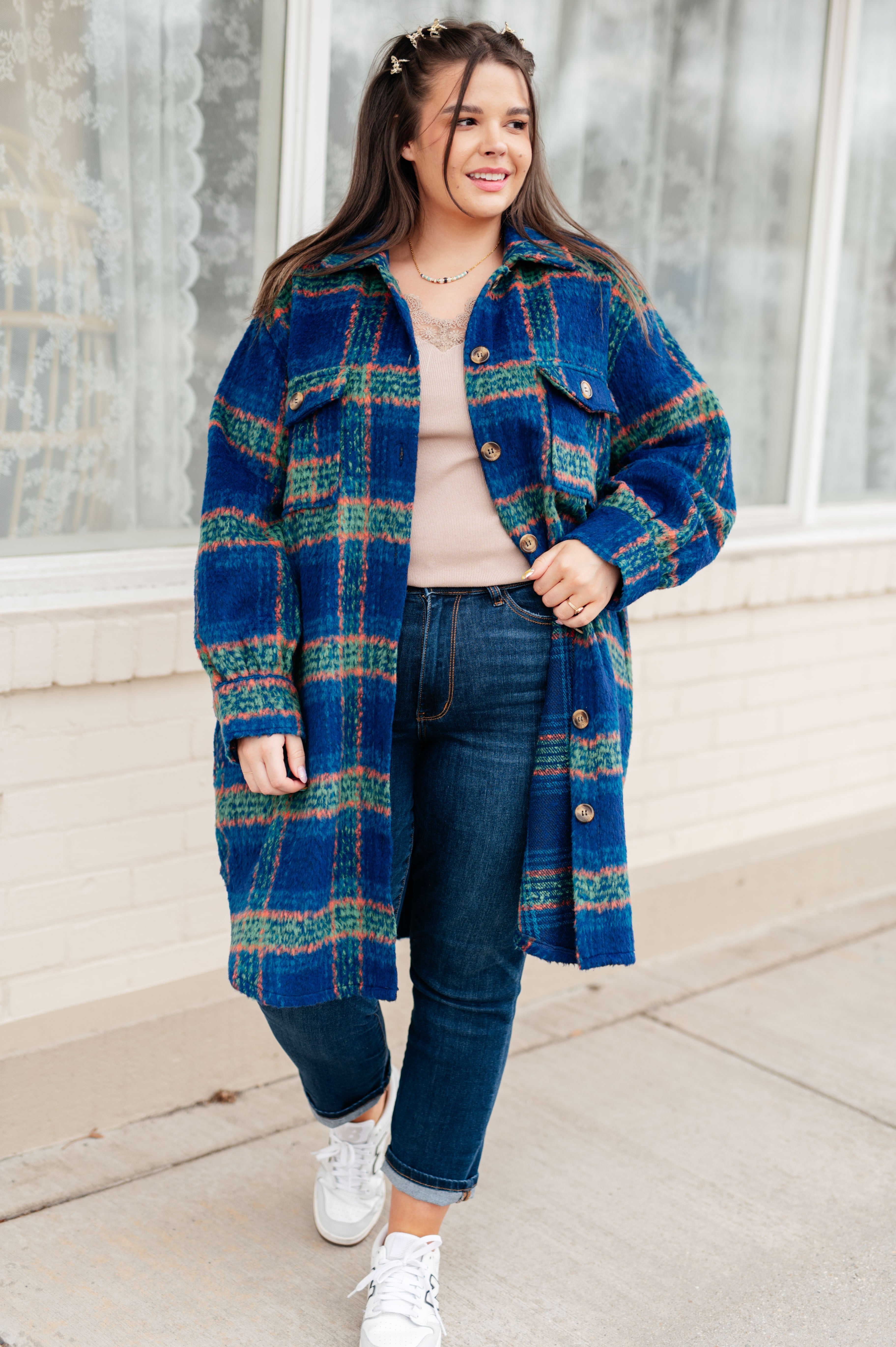 Woman smiling wearing a blue plaid shacket with green and orange stripes over a beige top and dark blue jeans, standing outdoors.