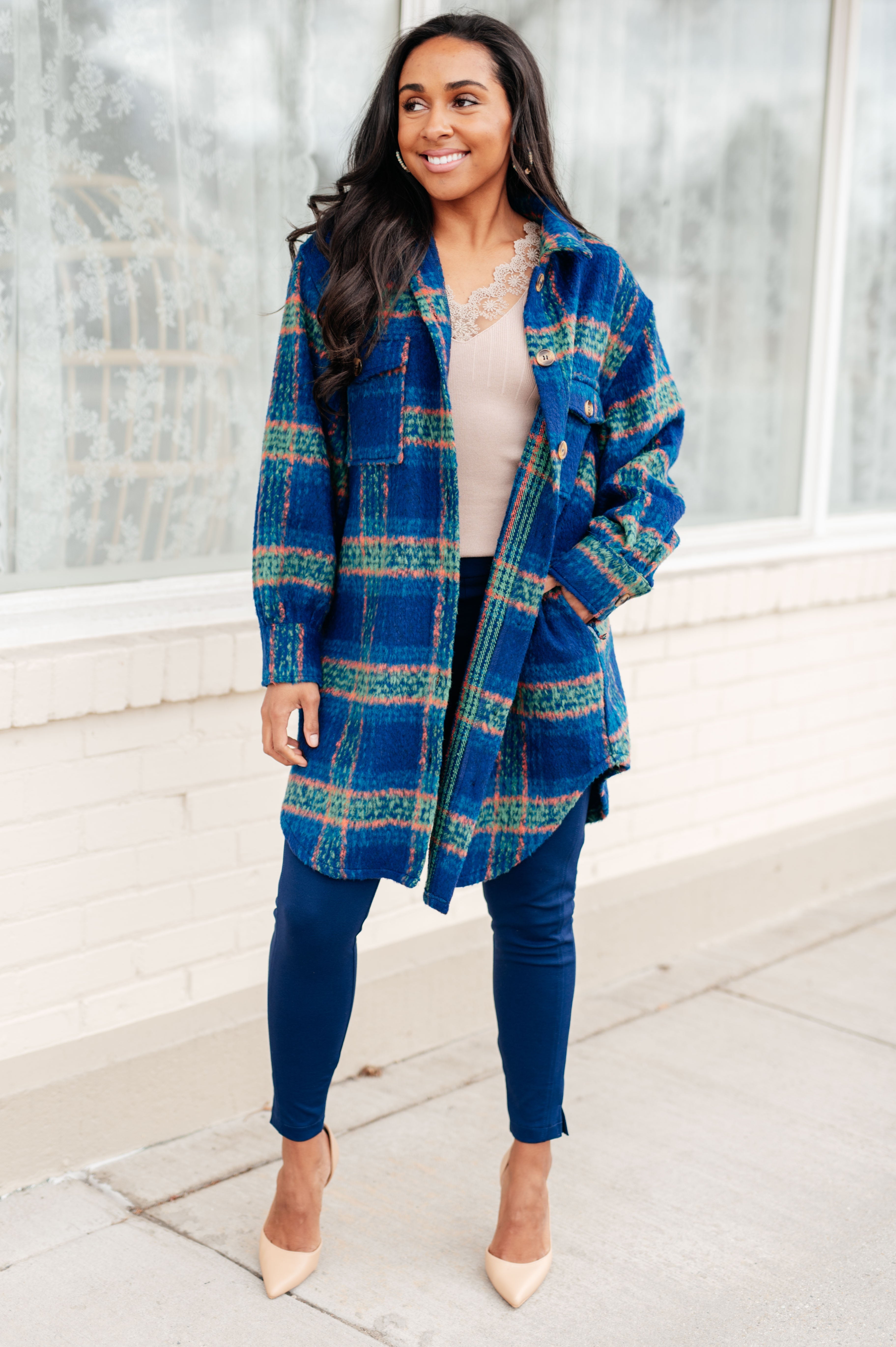 Woman wearing a blue plaid long coat with green and peach stripes over a light beige lace-trimmed top and navy pants, standing outdoors smiling.