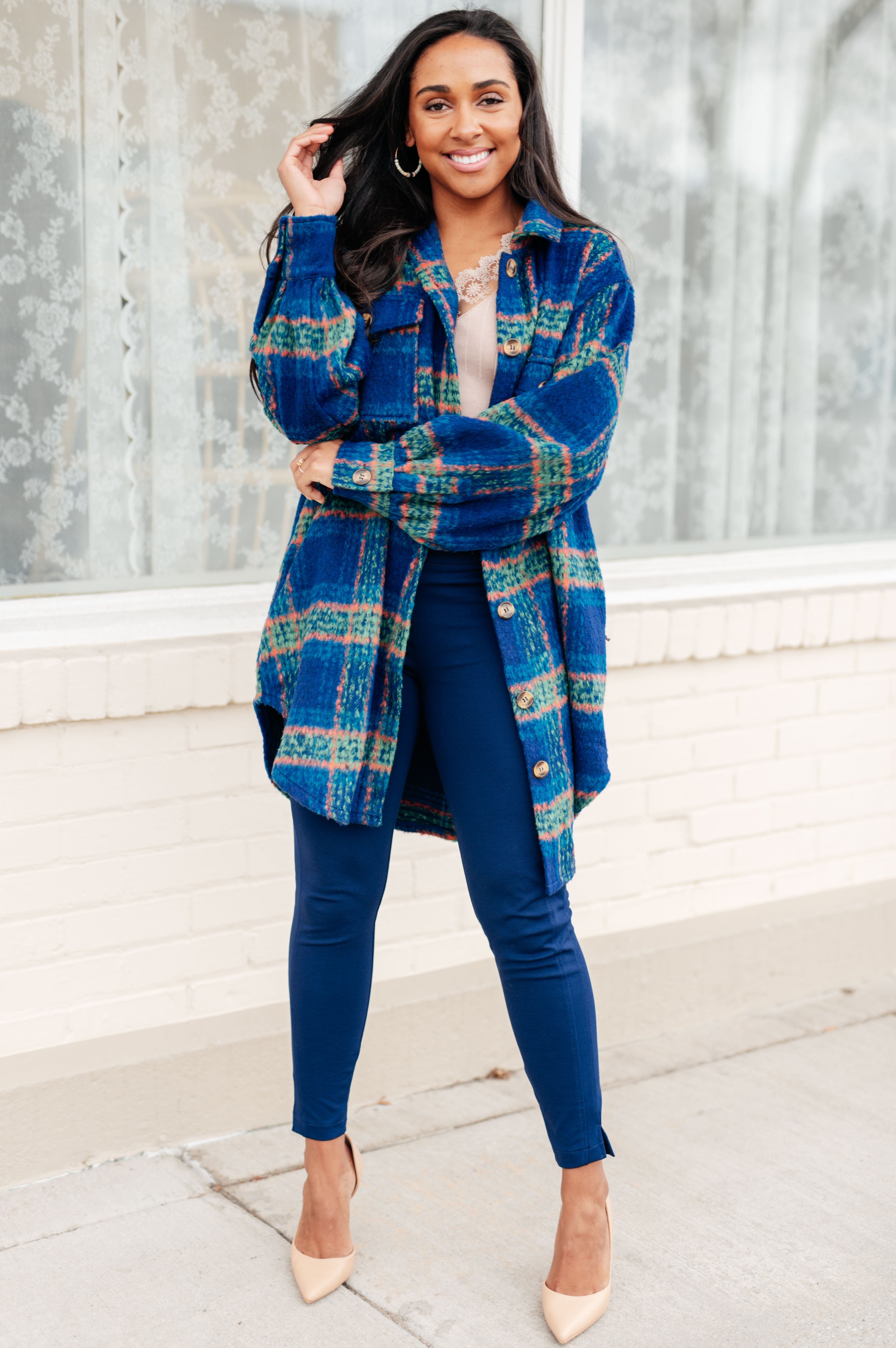 Smiling woman wearing a blue plaid long coat with green and orange stripes over a beige top and fitted dark blue pants, standing outside.