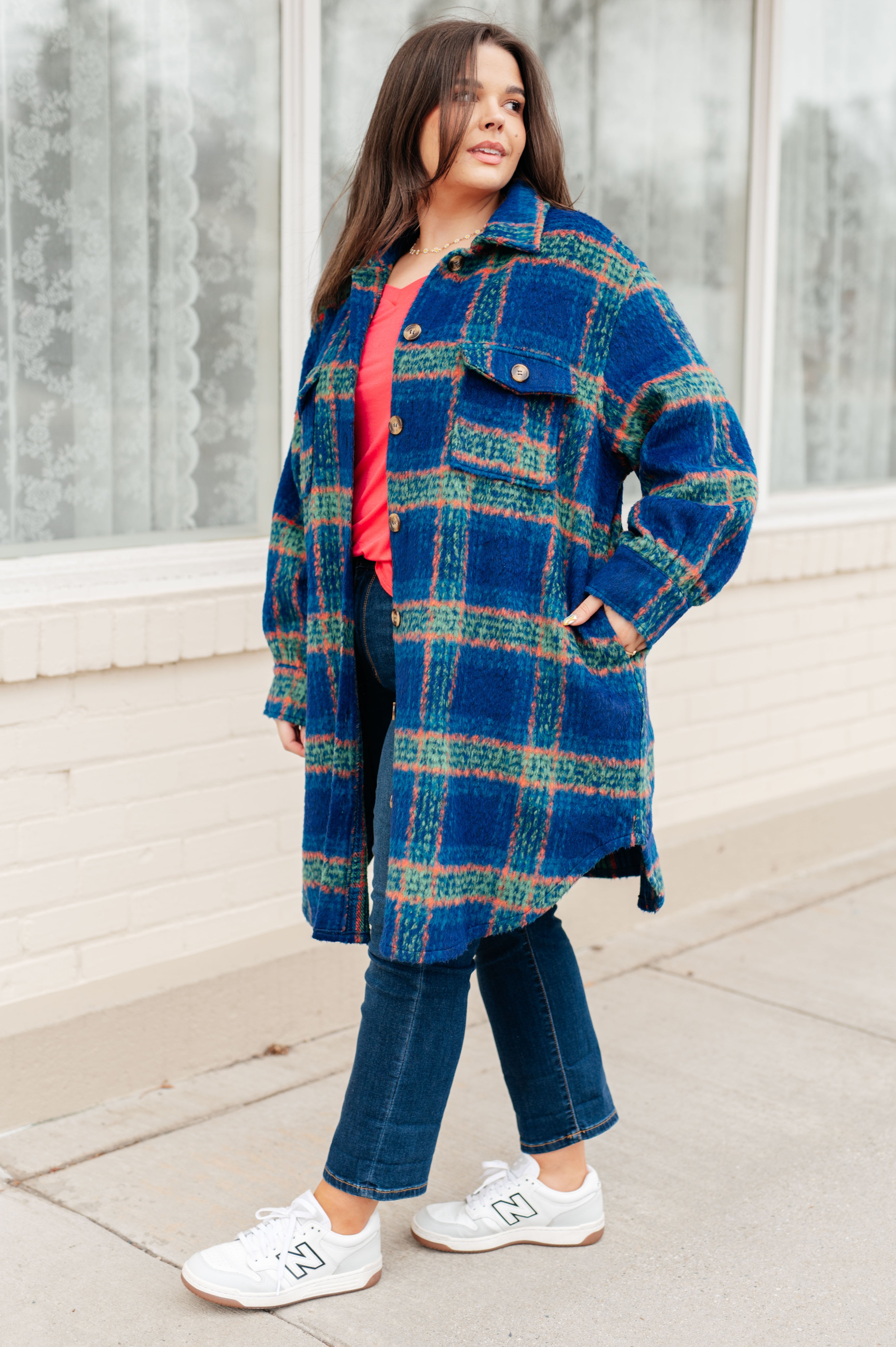 Woman wearing a cozy blue plaid coat with green and orange stripes over a coral top and cropped dark jeans, standing outdoors.