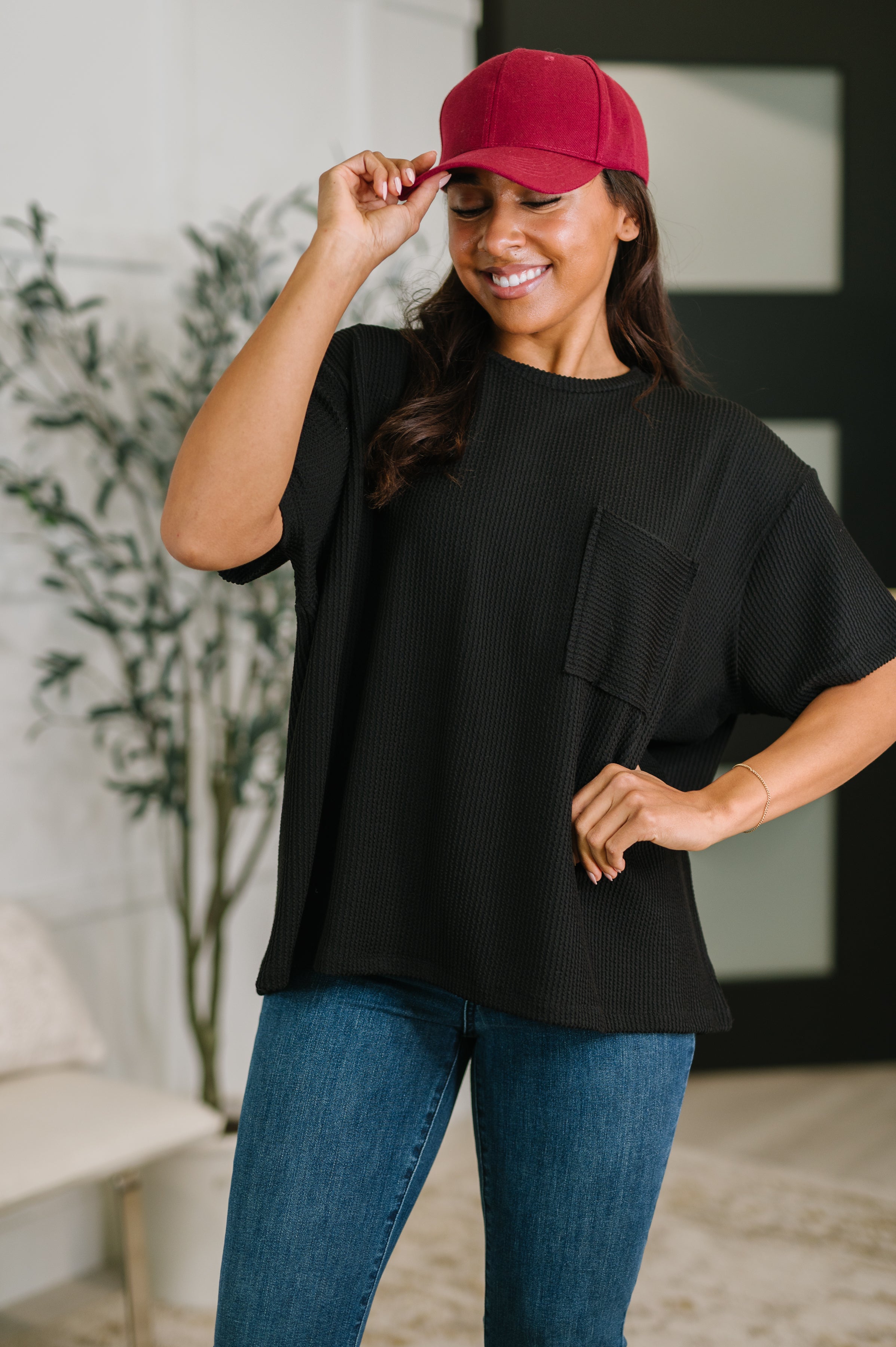 Woman wearing black textured boxy tee with chest pocket paired with blue jeans and red baseball cap indoors smiling