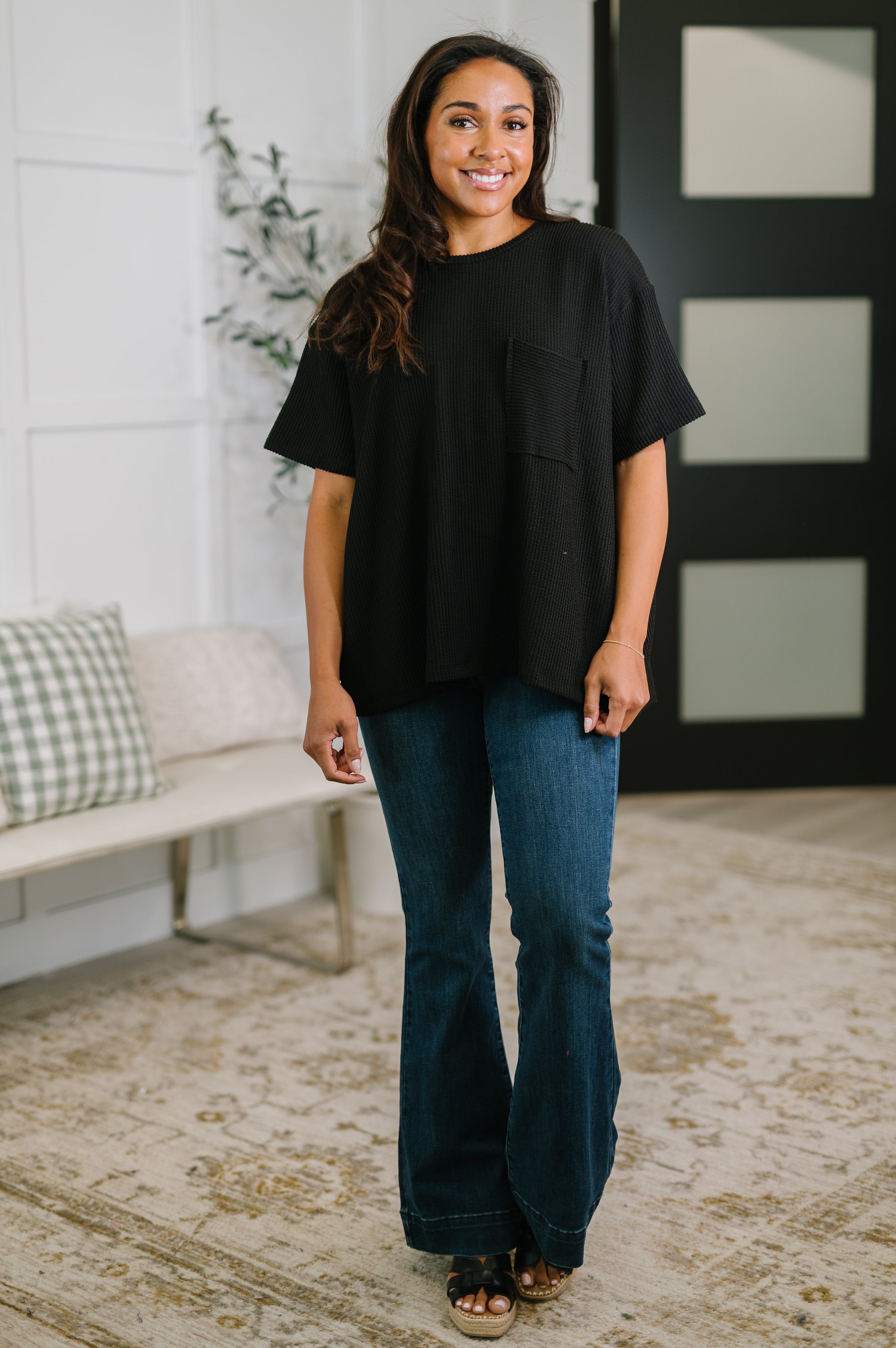 Woman smiling wearing black textured boxy tee with chest pocket and flared blue jeans in cozy indoor setting
