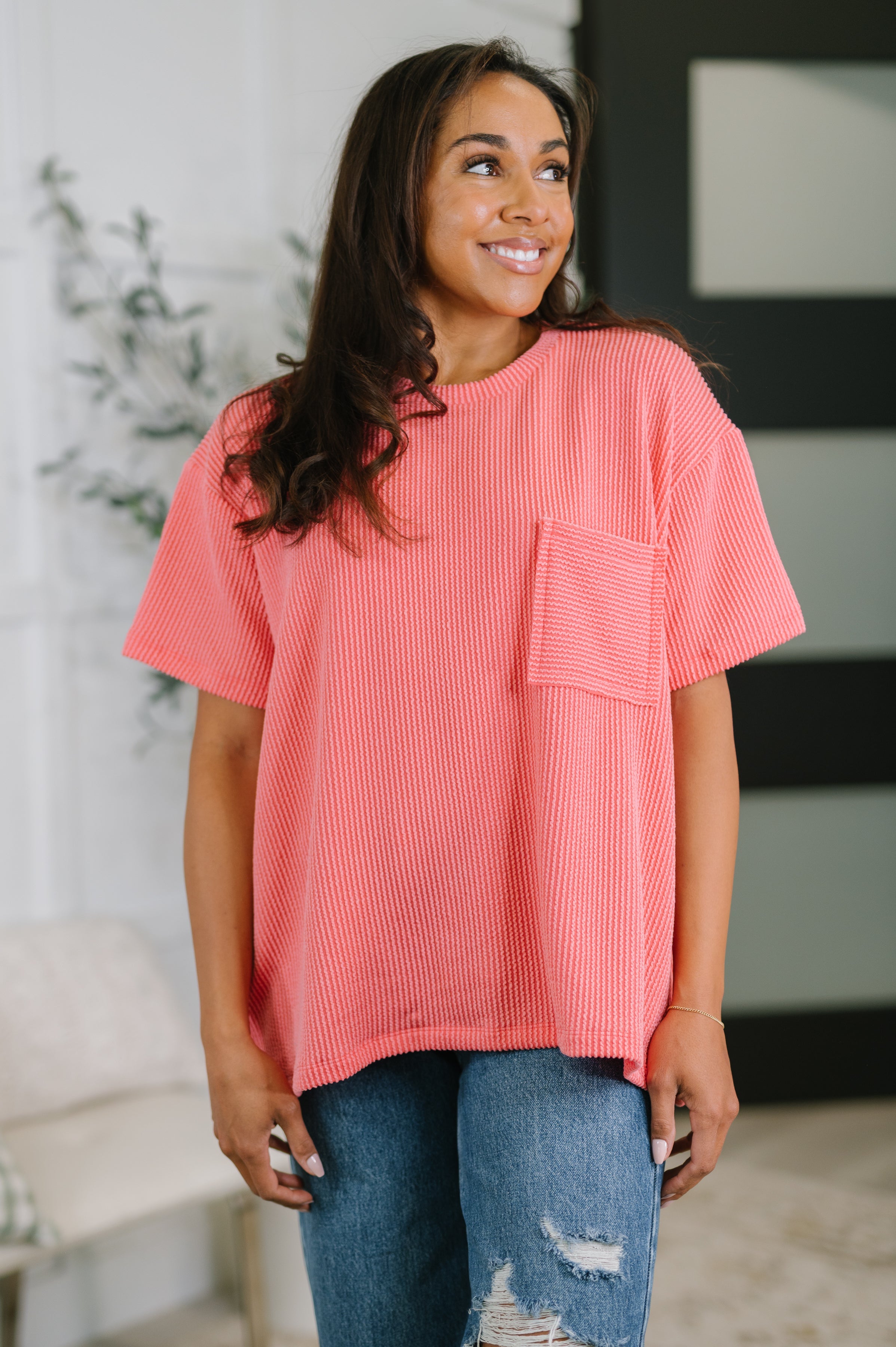 Smiling woman wearing salmon textured boxy tee with chest pocket and relaxed fit paired with ripped blue jeans indoors