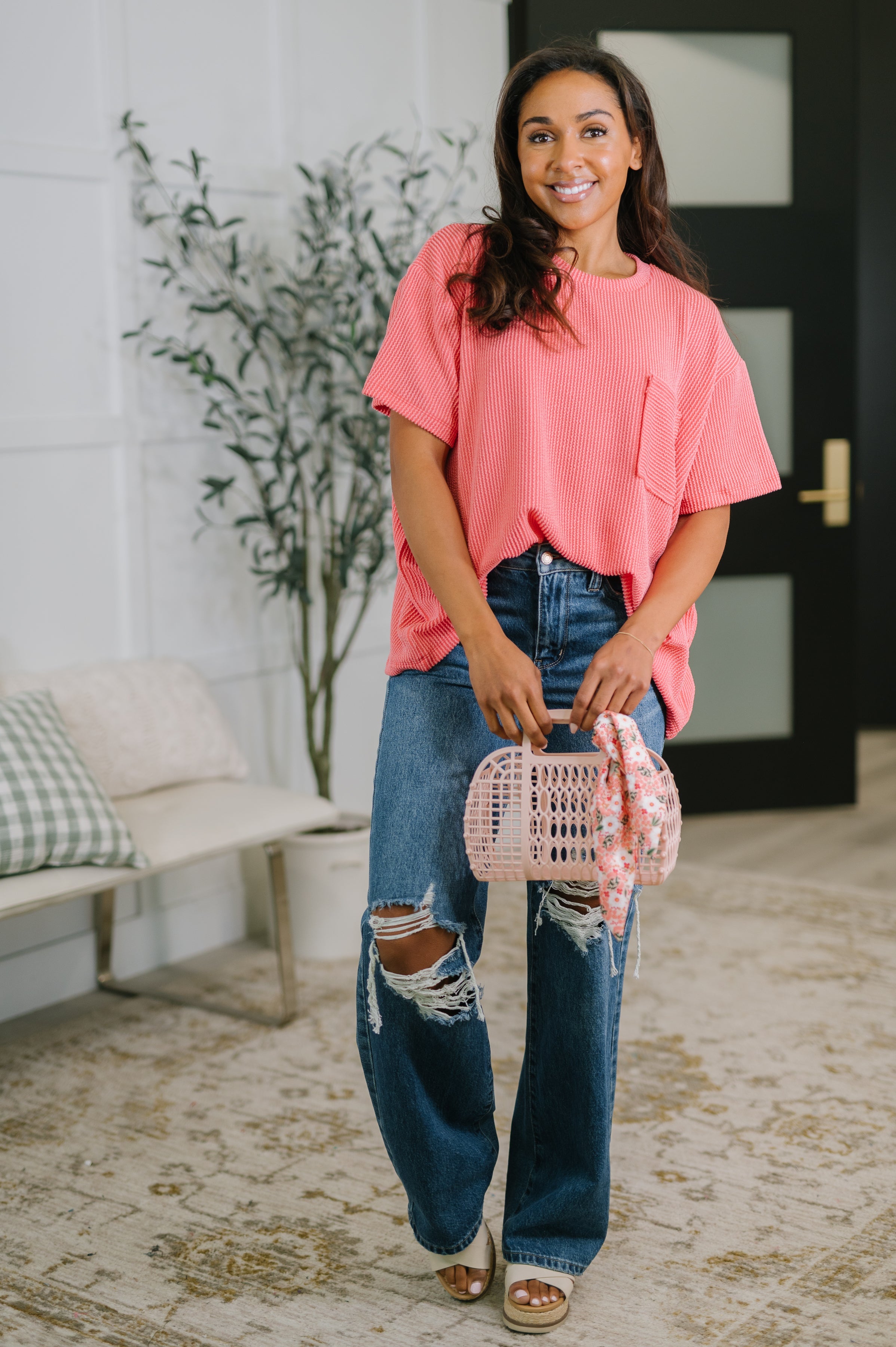 Woman wearing salmon textured boxy tee paired with ripped wide-leg jeans holding pink purse with floral scarf indoors