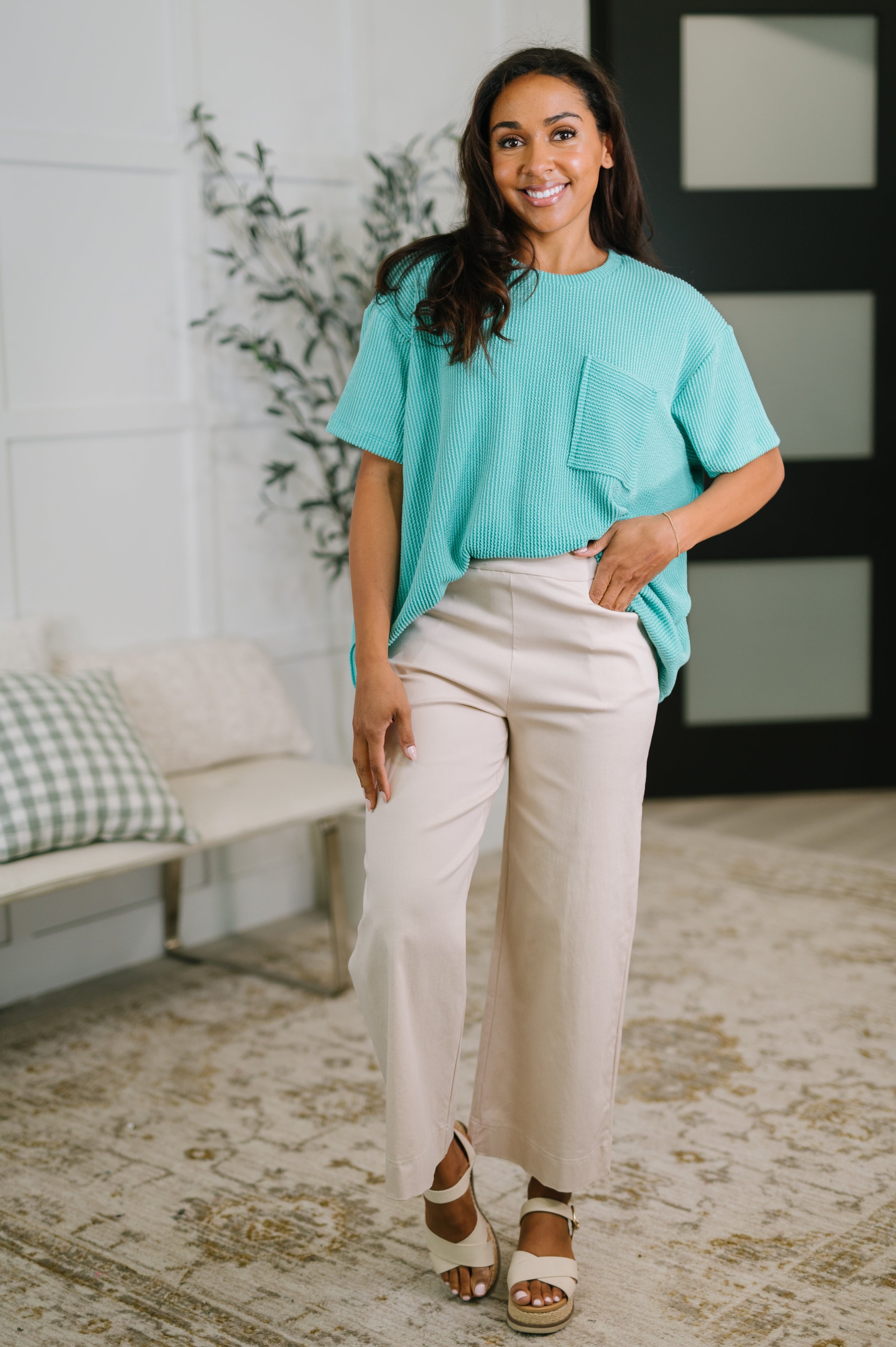 Woman wearing turquoise textured boxy tee with pocket paired with wide-leg beige pants and sandals indoors smiling