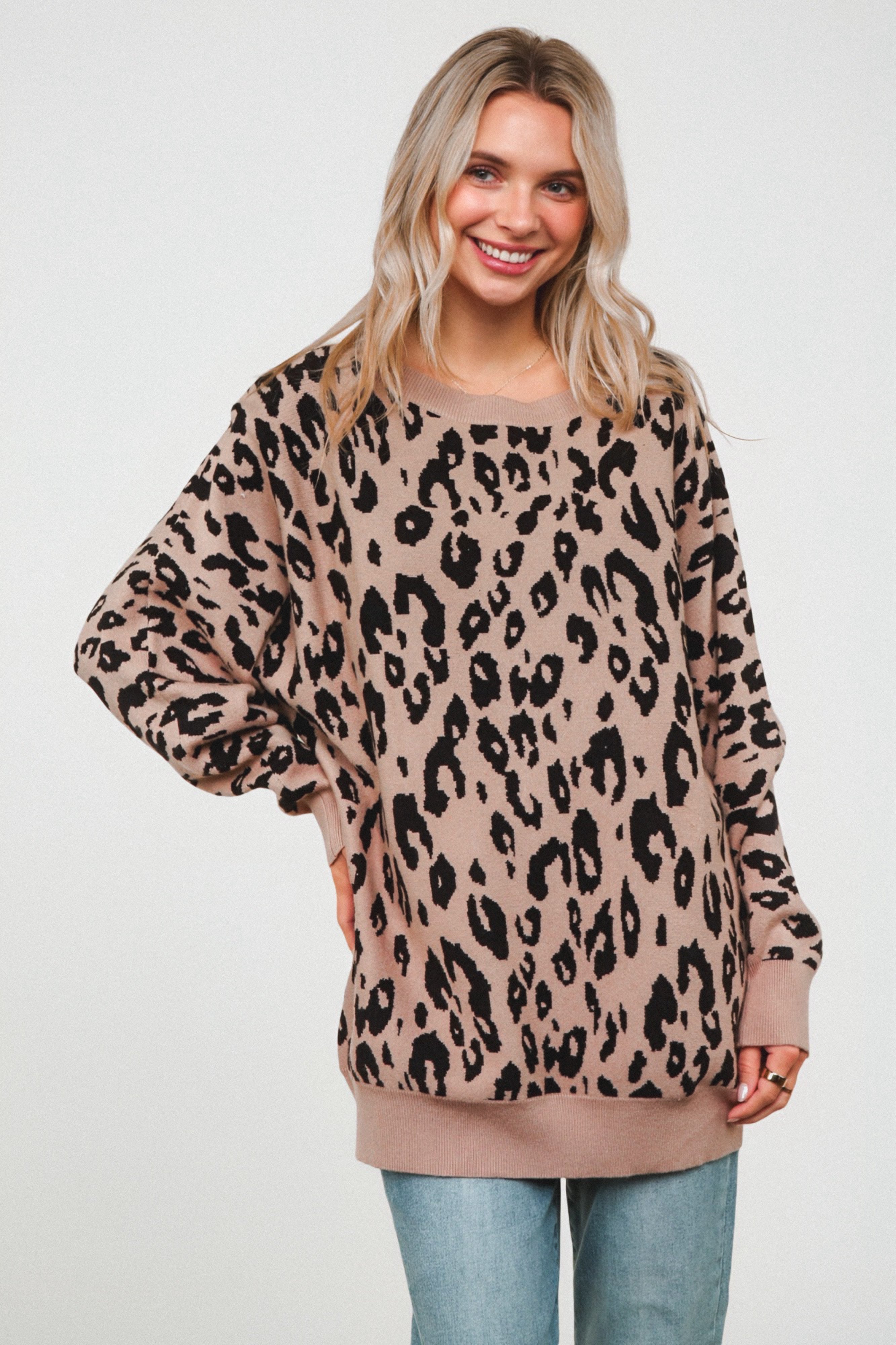 Woman smiling while wearing a relaxed mocha sweater featuring an all-over black leopard pattern, paired with light blue jeans against a plain background.