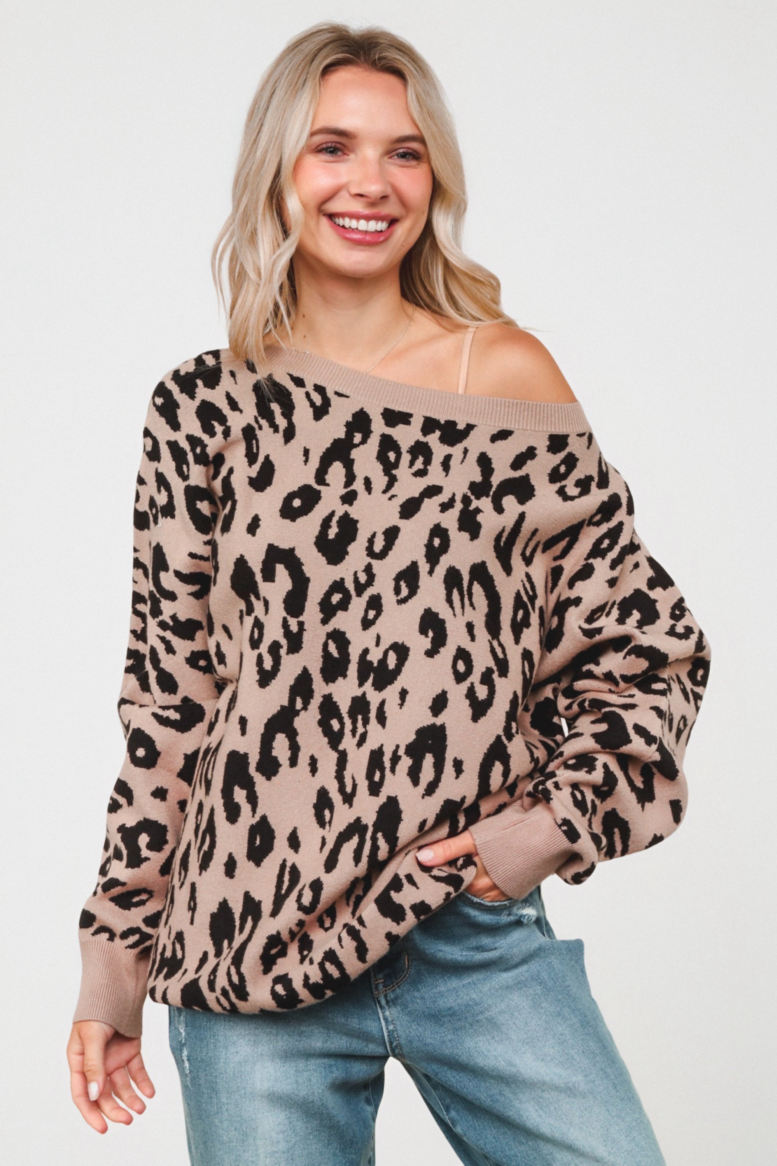 Smiling woman wearing a loose mocha sweater with a black leopard pattern, styled off one shoulder and paired with relaxed blue jeans.