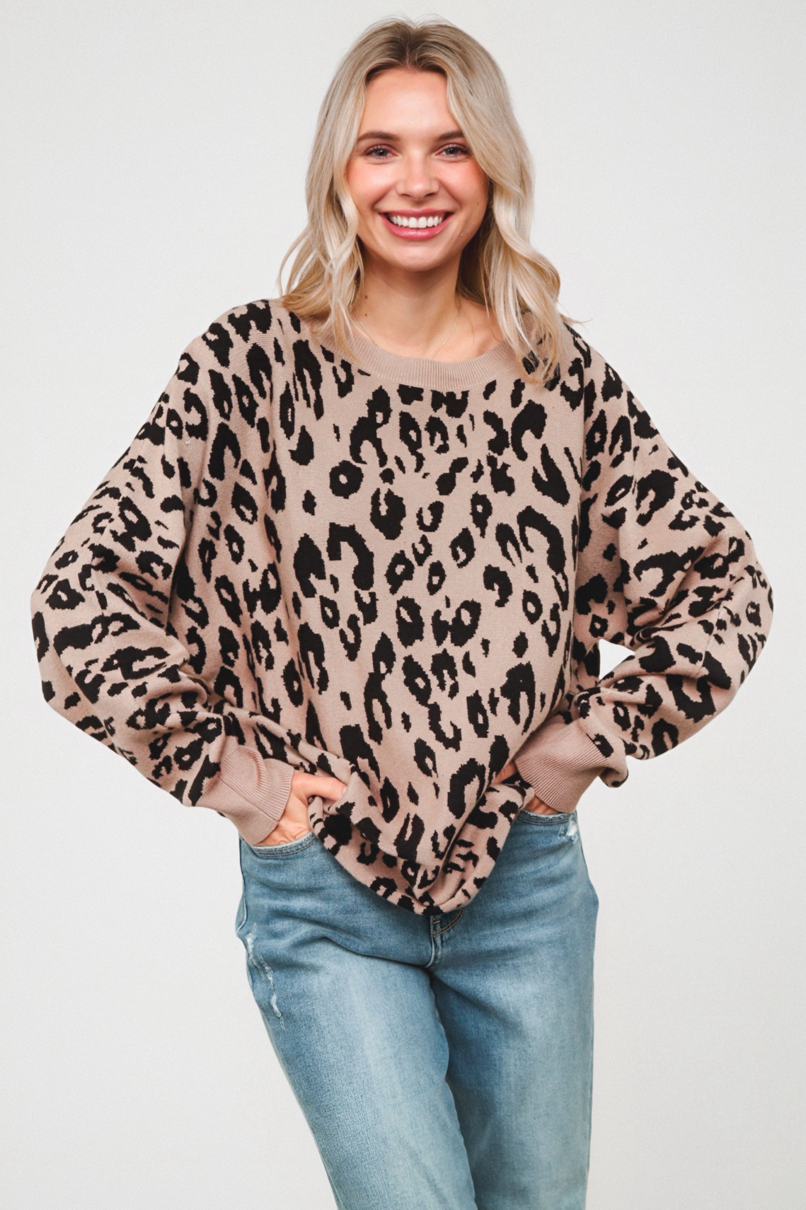 Woman smiling while wearing a relaxed mocha sweater with a bold black leopard pattern, paired with light blue jeans and standing against a plain background.