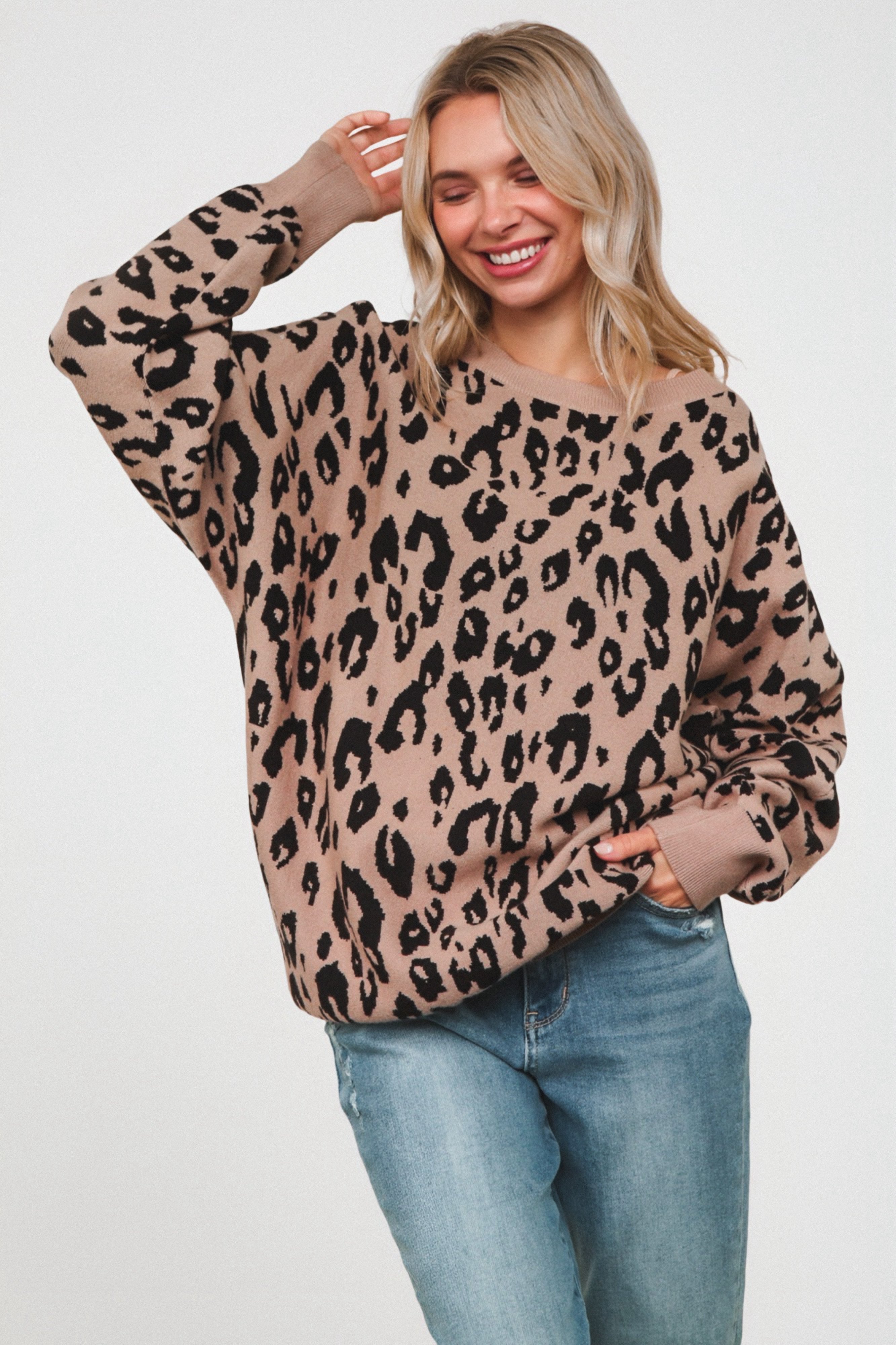 Smiling woman wearing a loose mocha sweater with bold black leopard print, paired with light blue jeans, standing against a plain background.