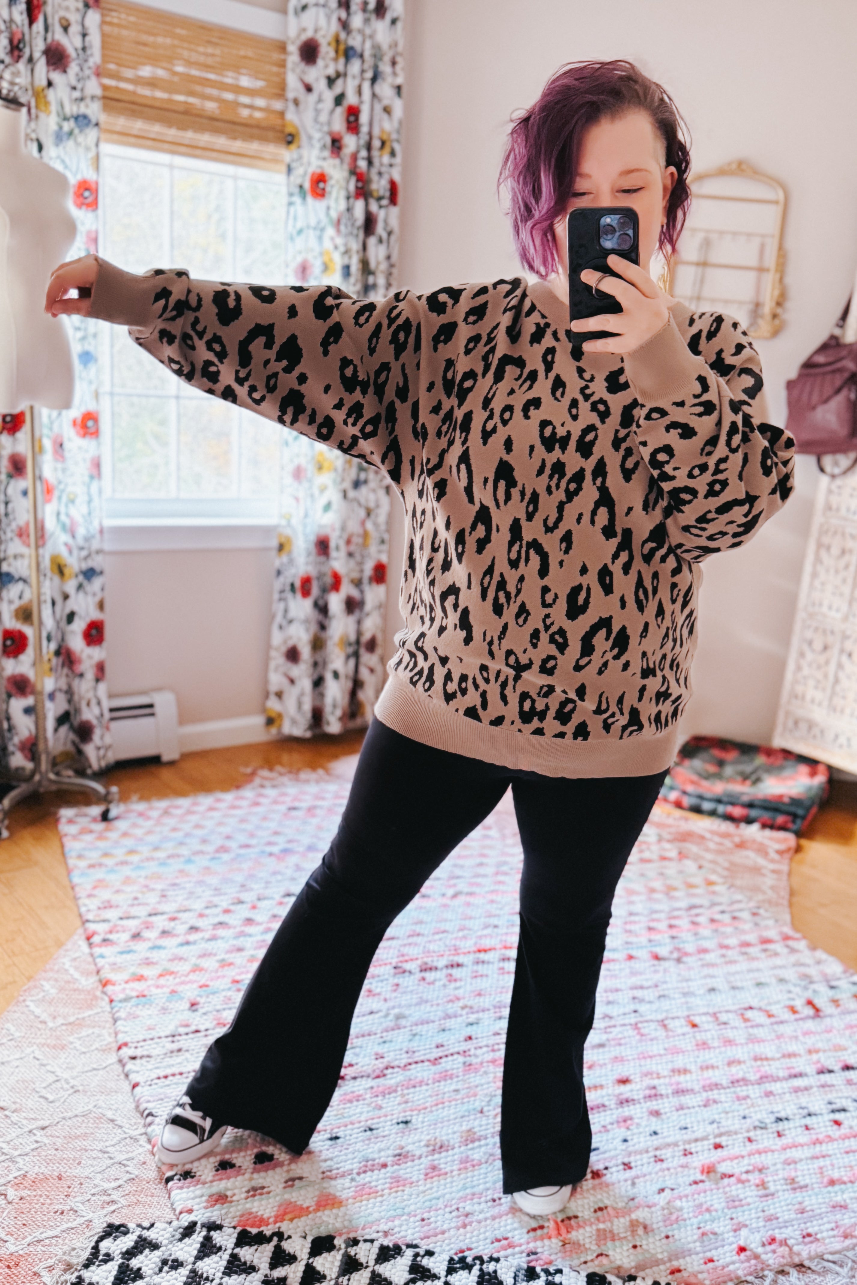 Woman taking a selfie wearing a mocha sweater with a black leopard print, paired with black flared pants and sneakers in a cozy room.