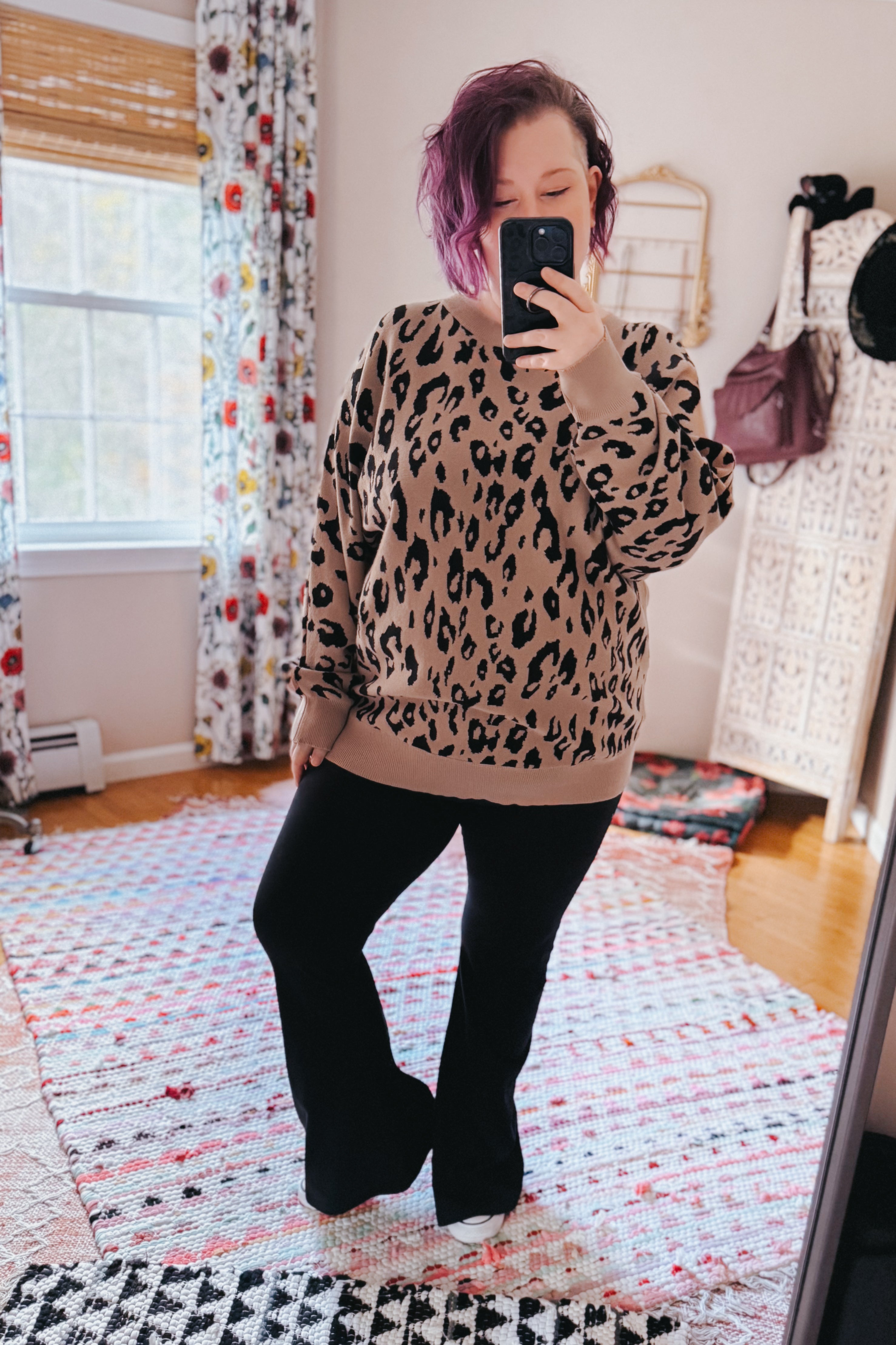Woman taking a mirror selfie wearing a relaxed mocha sweater with a black leopard print, paired with black pants in a cozy room.