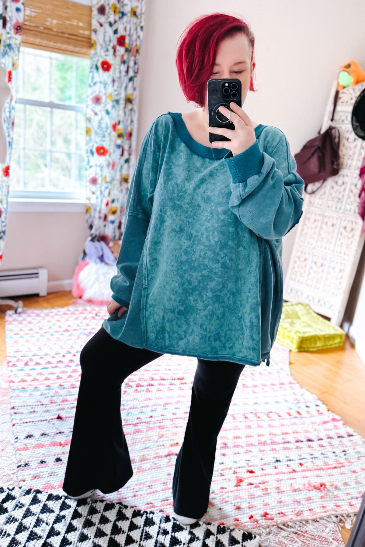Red haired woman models a blue mineral wash pullover with oversized sleeves and black flared pants in a colorful bedroom mirror selfie