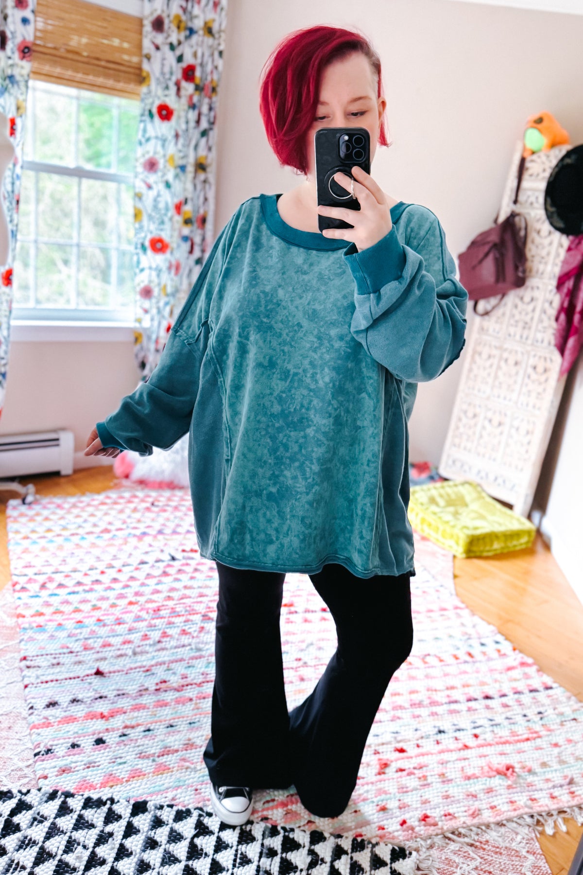 Woman with short red hair takes a bedroom mirror selfie wearing a blue mineral wash pullover black flared pants and white sneakers