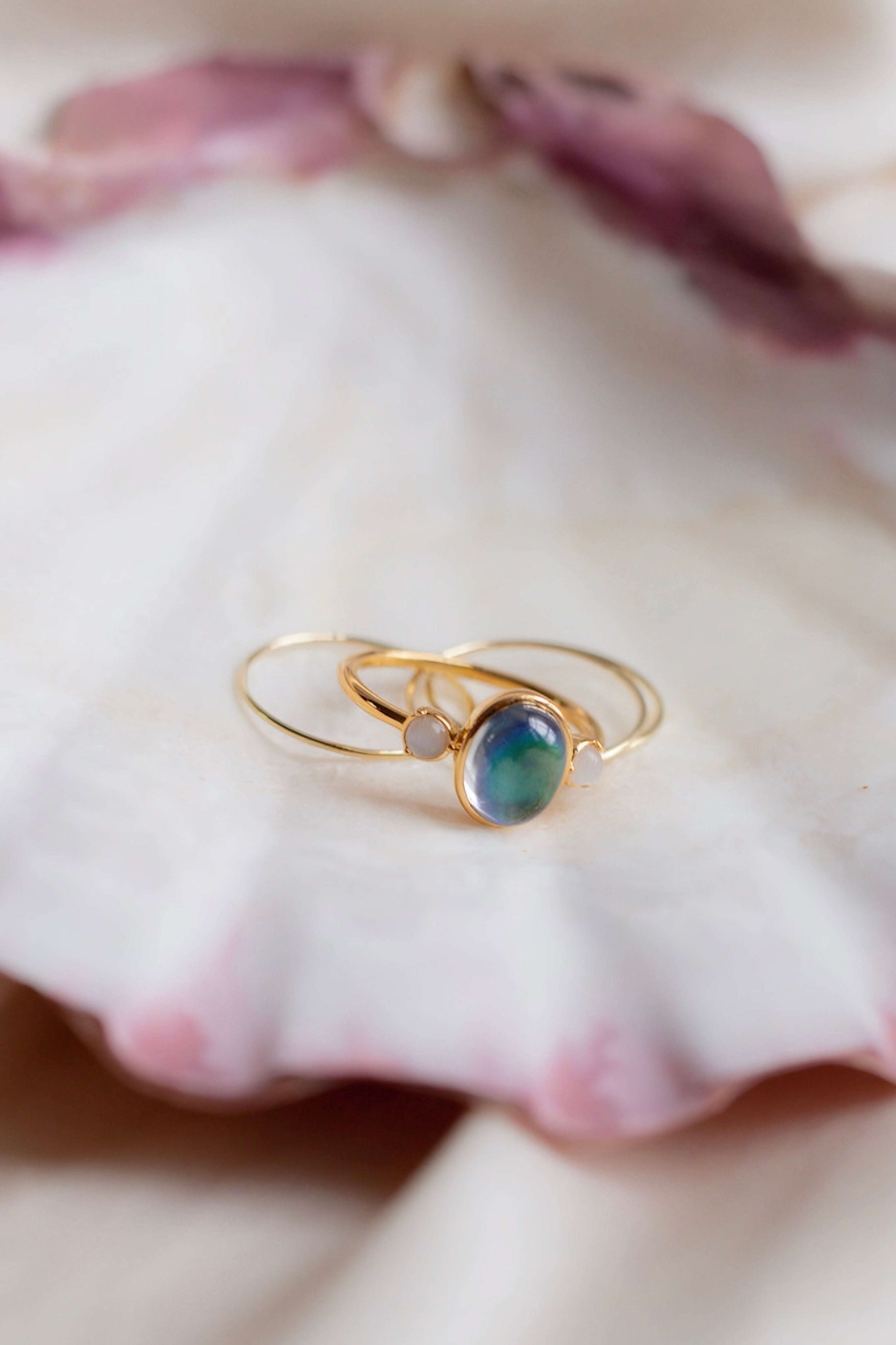 Delicate gold mood ring with blue-green stone and two slim gold bands resting on a soft pink-edged shell surface