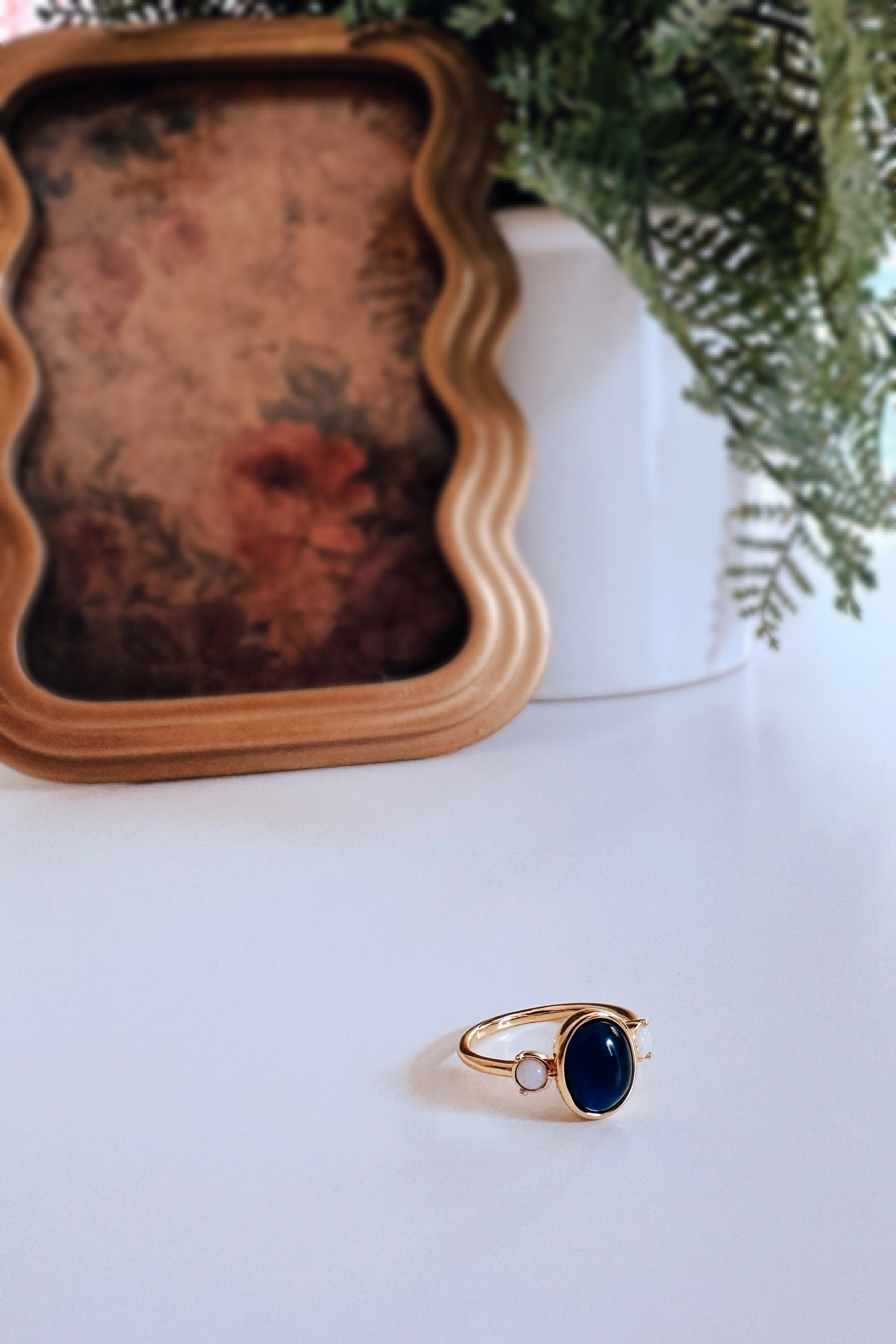 Gold Odette Bejeweled Mood Ring with deep blue oval stone and small pale round stone beside plant and vintage frame