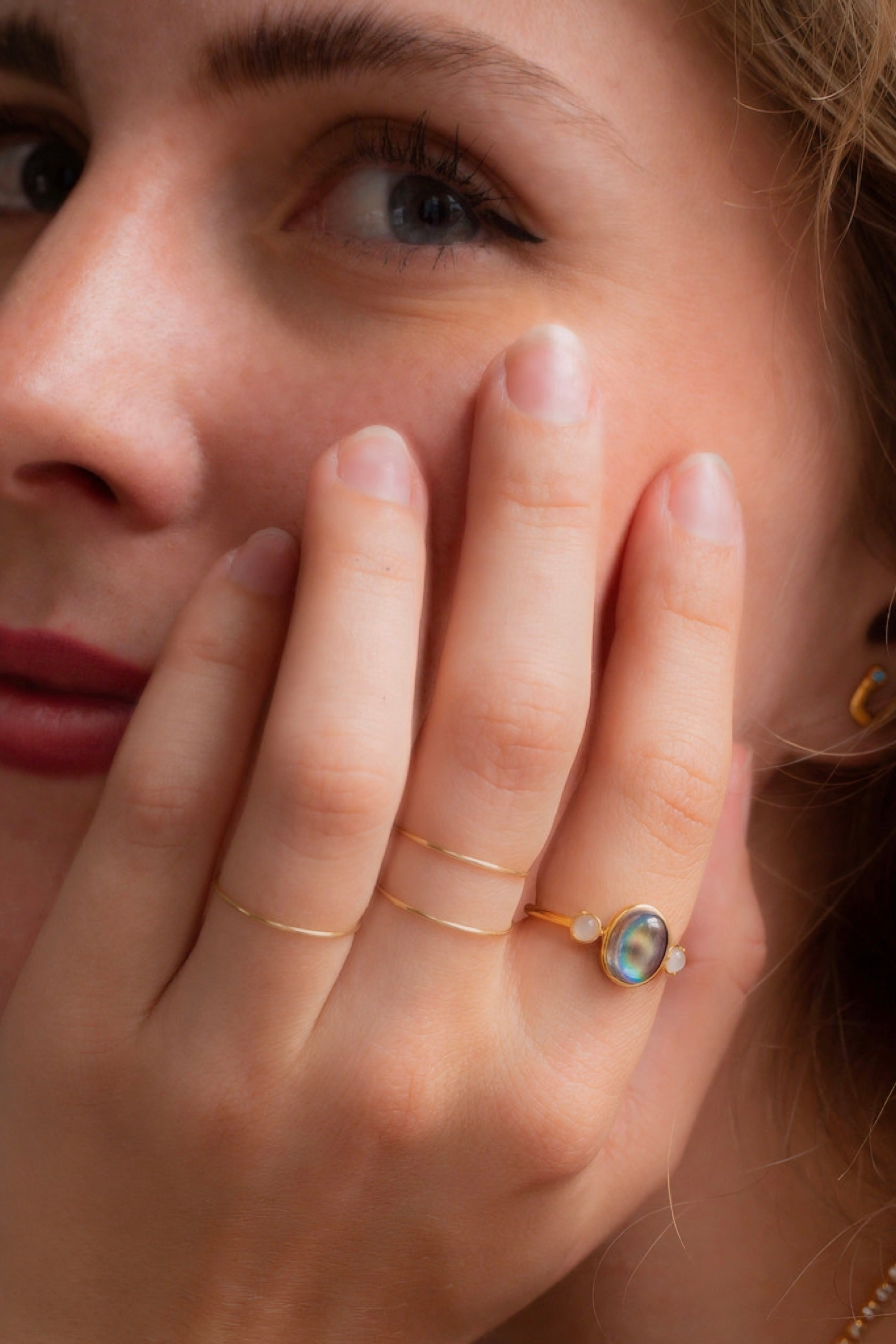 Hand wearing gold Odette Bejeweled Mood Ring with iridescent oval stone and delicate bands near face with natural makeup