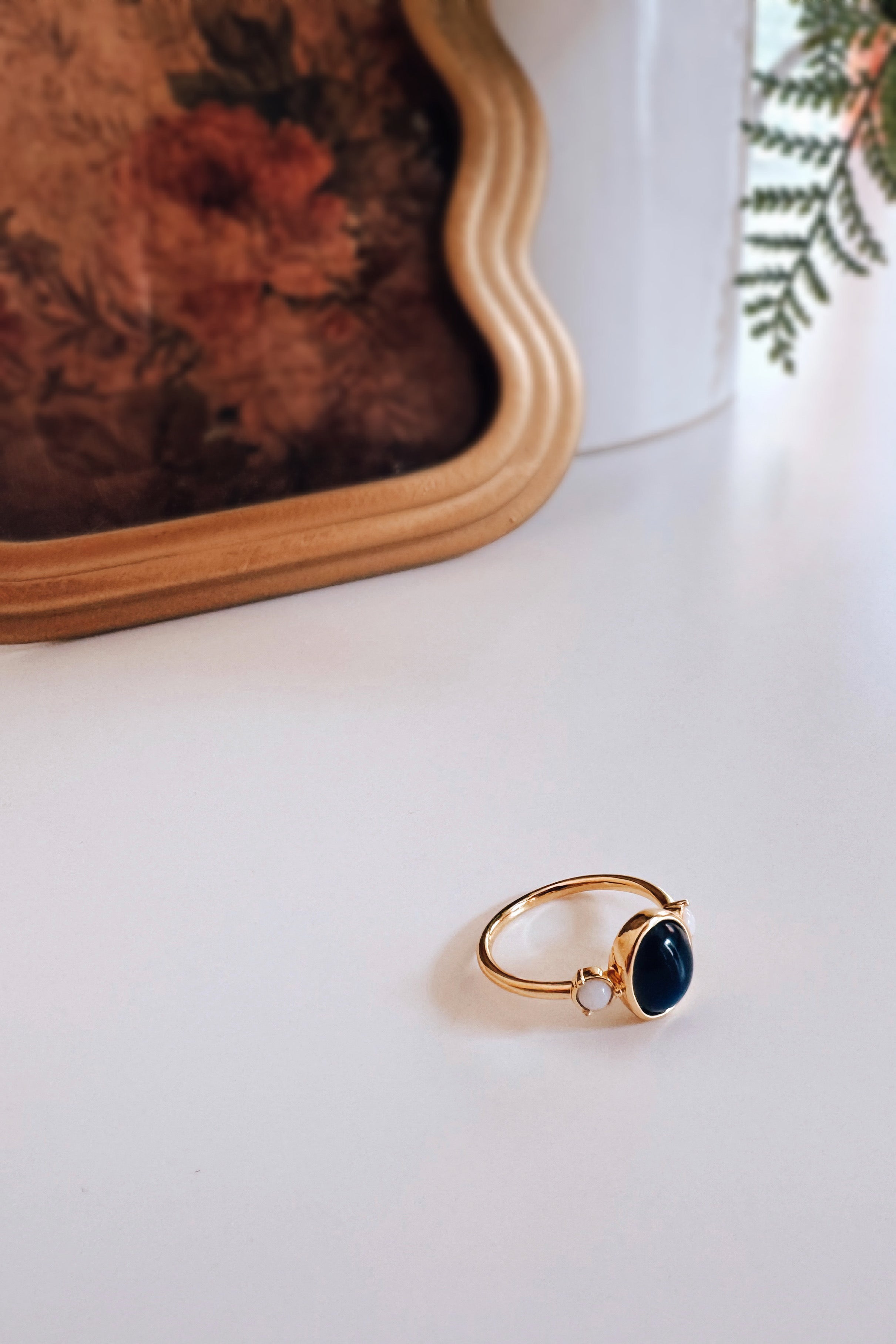 Gold-tone ring featuring a dark oval stone and small pale round stone on minimalist white surface with plant shadows
