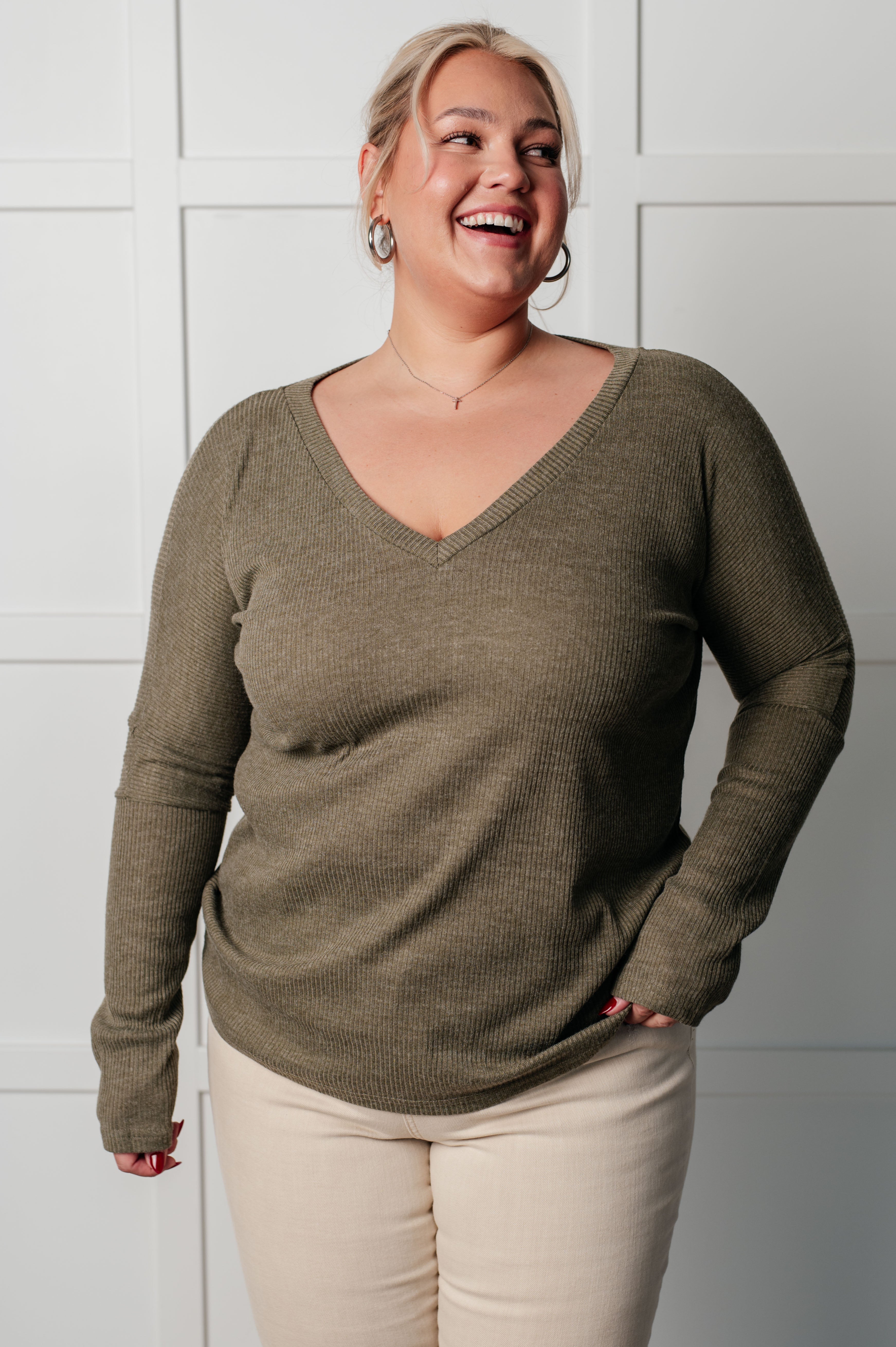 Plus-size woman smiling wearing olive ribbed knit V-neck long sleeve top and light pants against white paneled wall