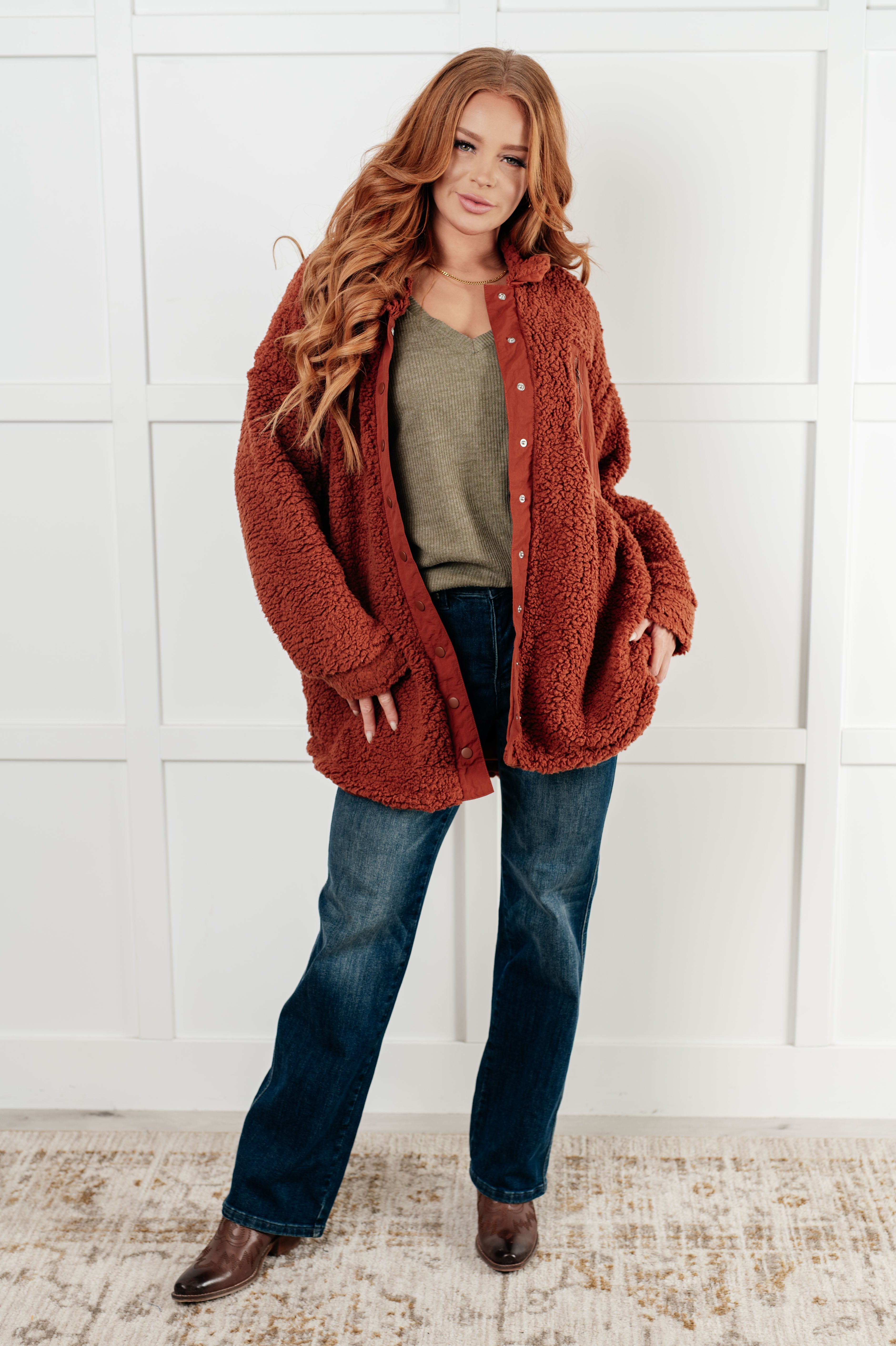 Woman with long red hair wearing olive ribbed V-neck top, oversized rust sherpa jacket, dark jeans, and brown boots indoors