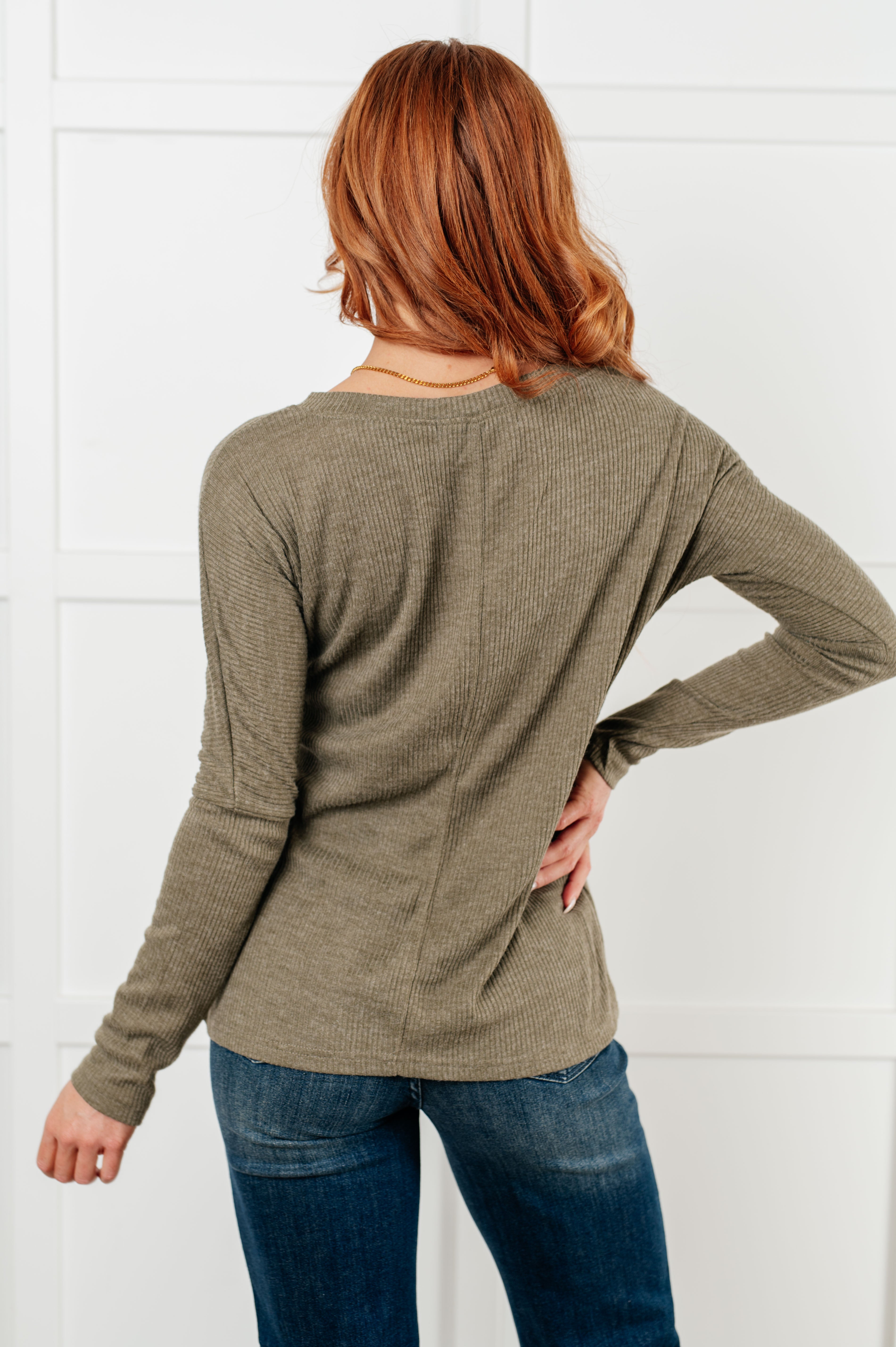 Red-haired woman modeling olive ribbed knit long sleeve top viewed from back with hand on hip and blue jeans