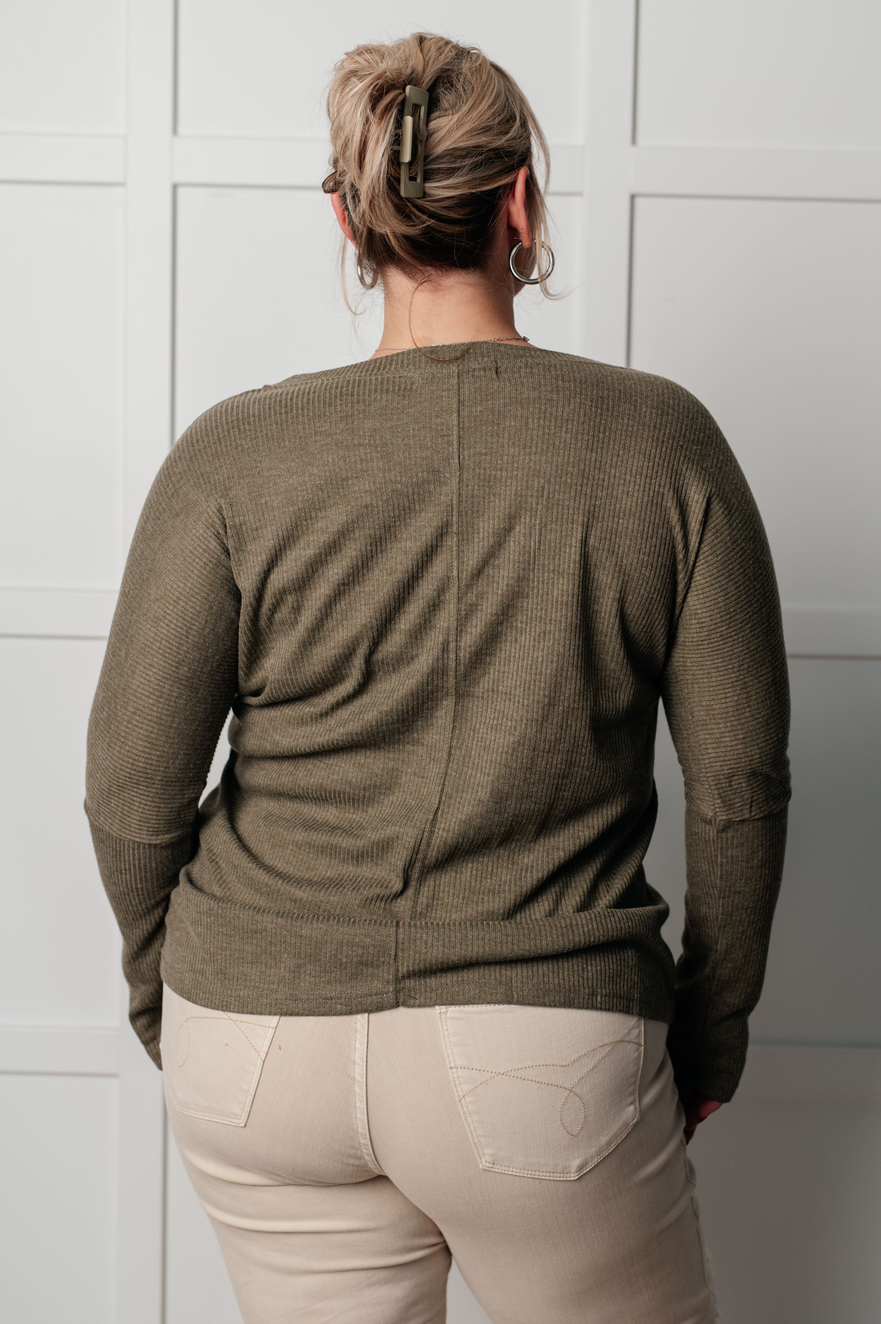Plus-size woman with blond hair wearing olive ribbed knit long sleeve top and beige pants against white paneled wall