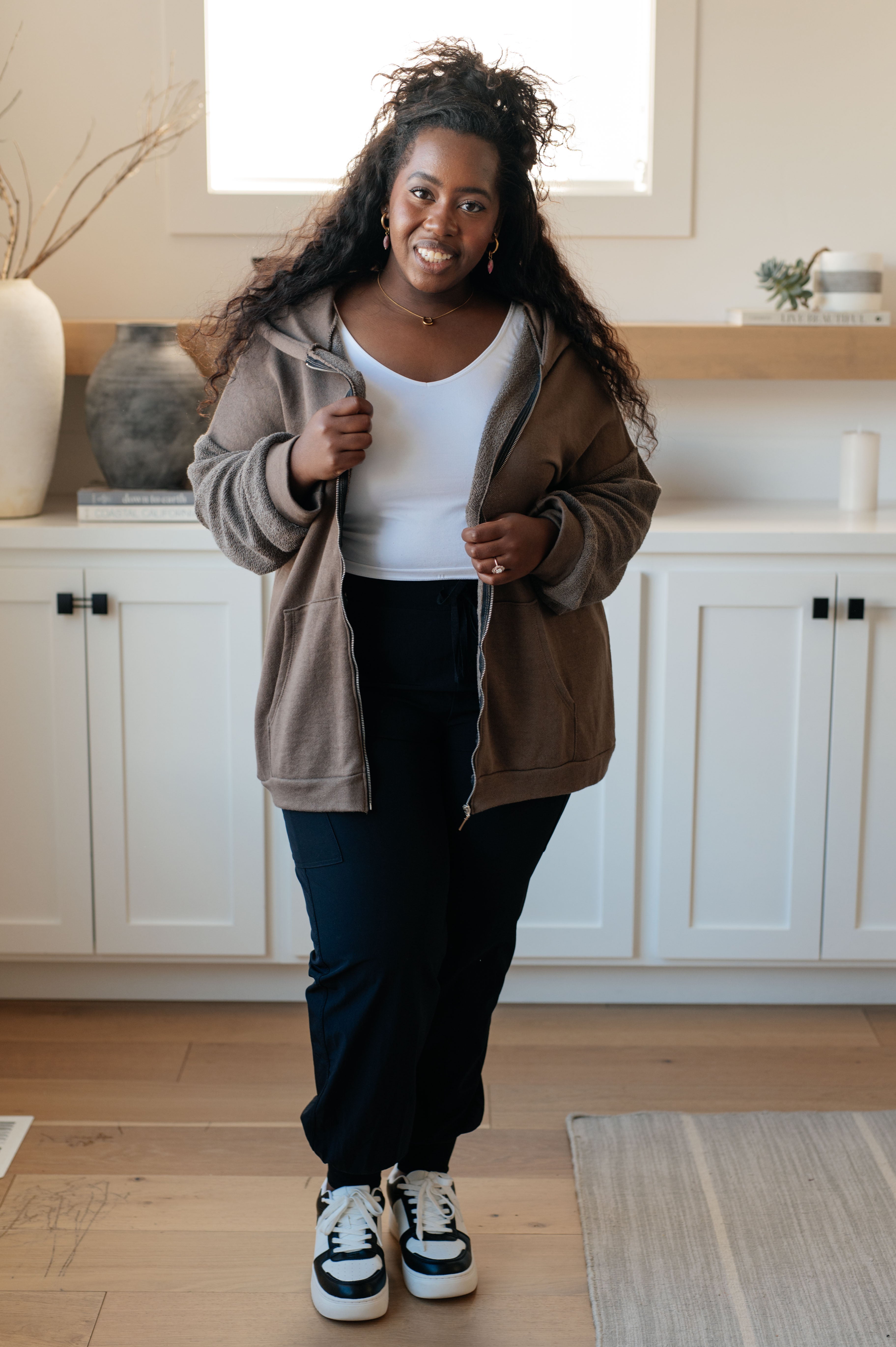 Woman wearing a brown zip-up hoodie over a white tank top over black jeans in a casual indoor setting.