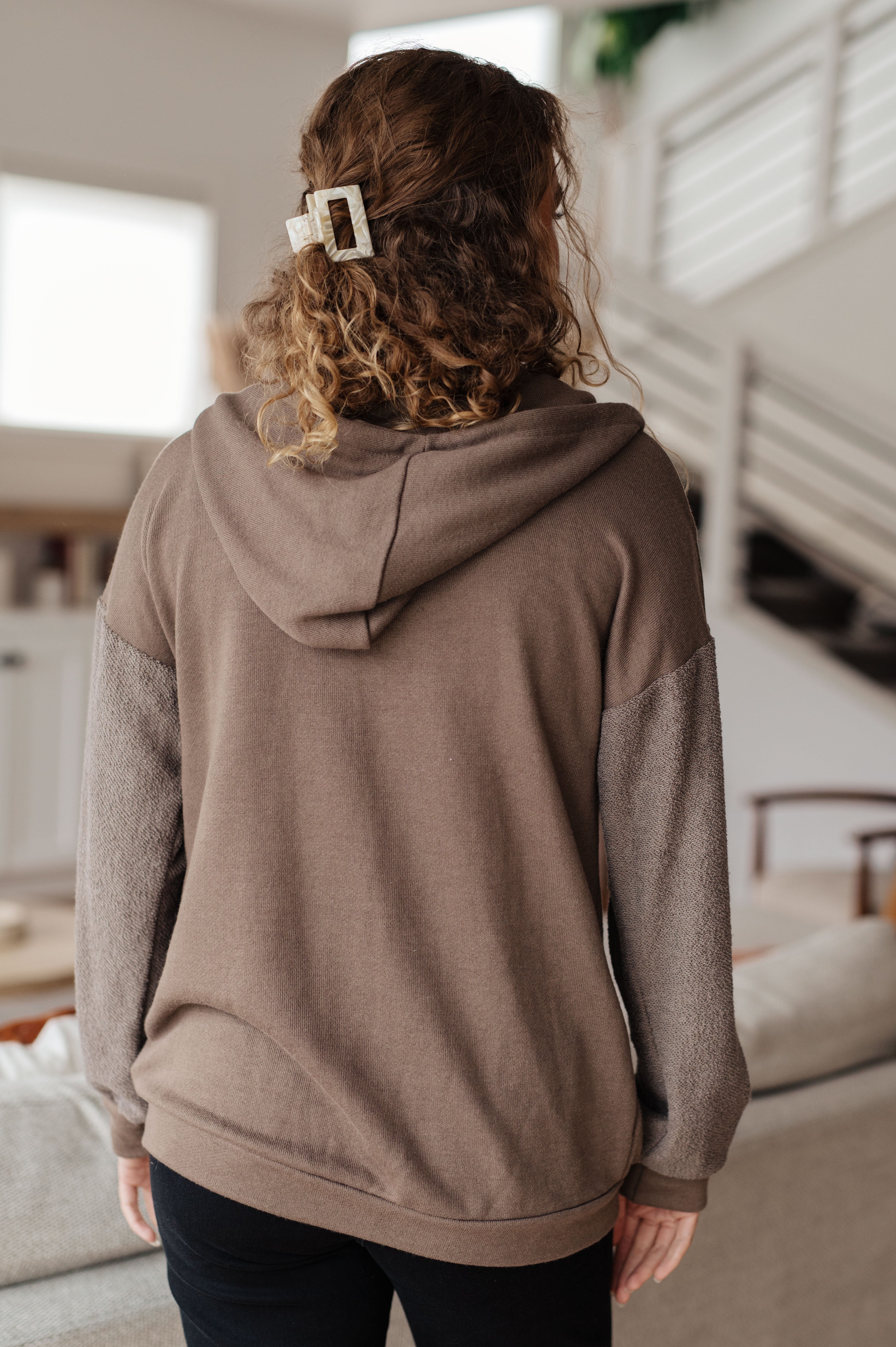 Woman with her back facing the camera wearing a brown zip-up hoodie over over black jeans in a casual indoor setting.