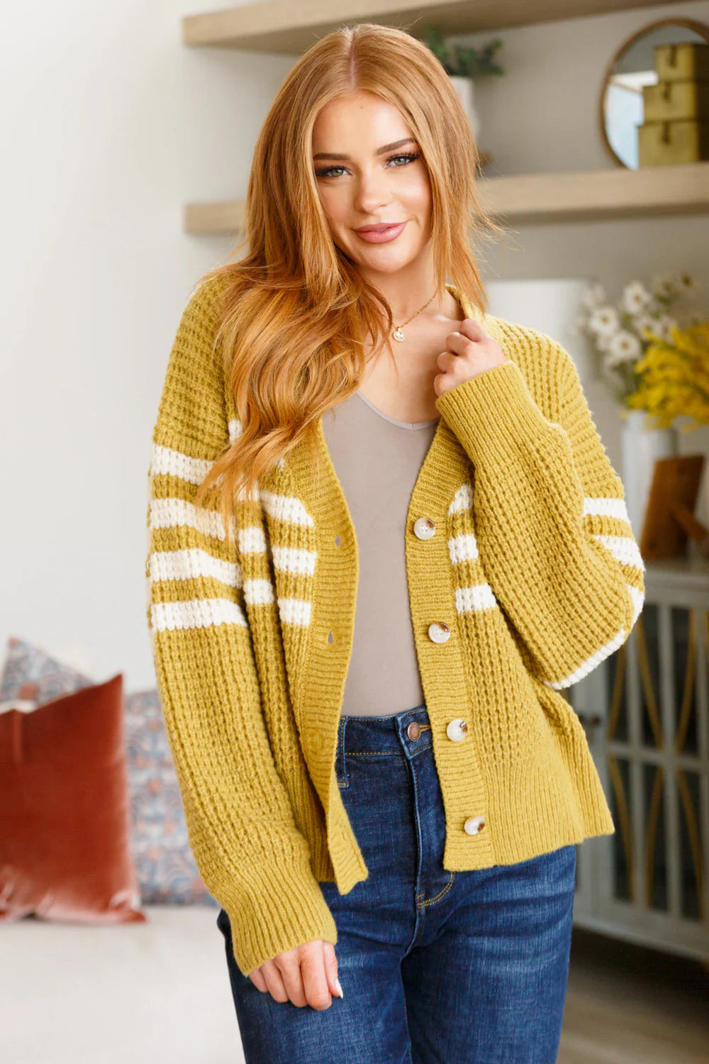 Woman wearing mustard knit cardigan with white stripes and blue jeans in bright cozy room with plants and shelves
