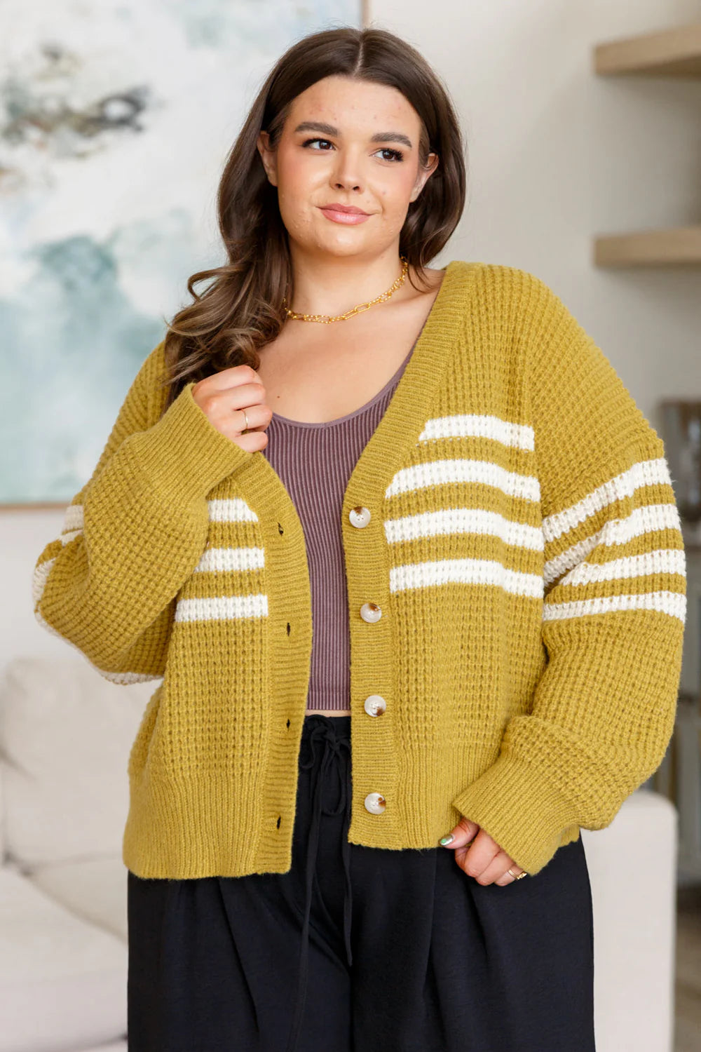 Plus-size woman wearing mustard knit cardigan with white stripes and black pants in relaxed indoor setting
