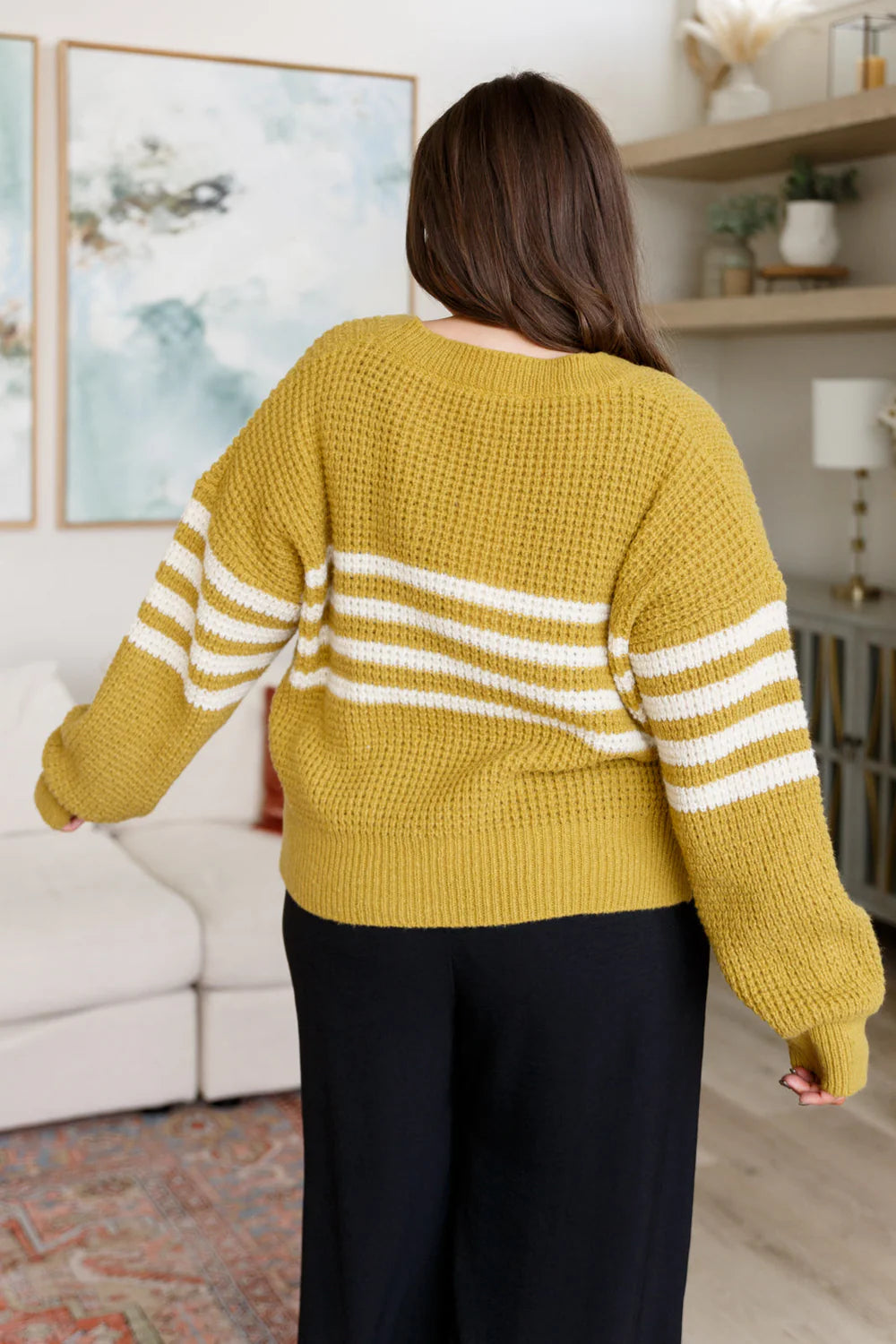 Woman seen from behind wearing mustard and white striped knit cardigan with black pants in cozy living room setting