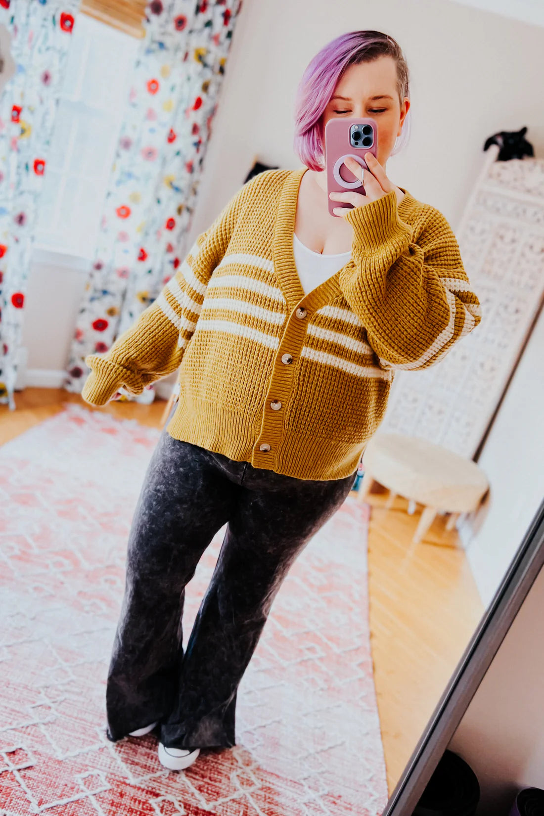 Woman with lavender hair wearing mustard cardigan with white stripes and dark flared pants taking a mirror selfie indoors