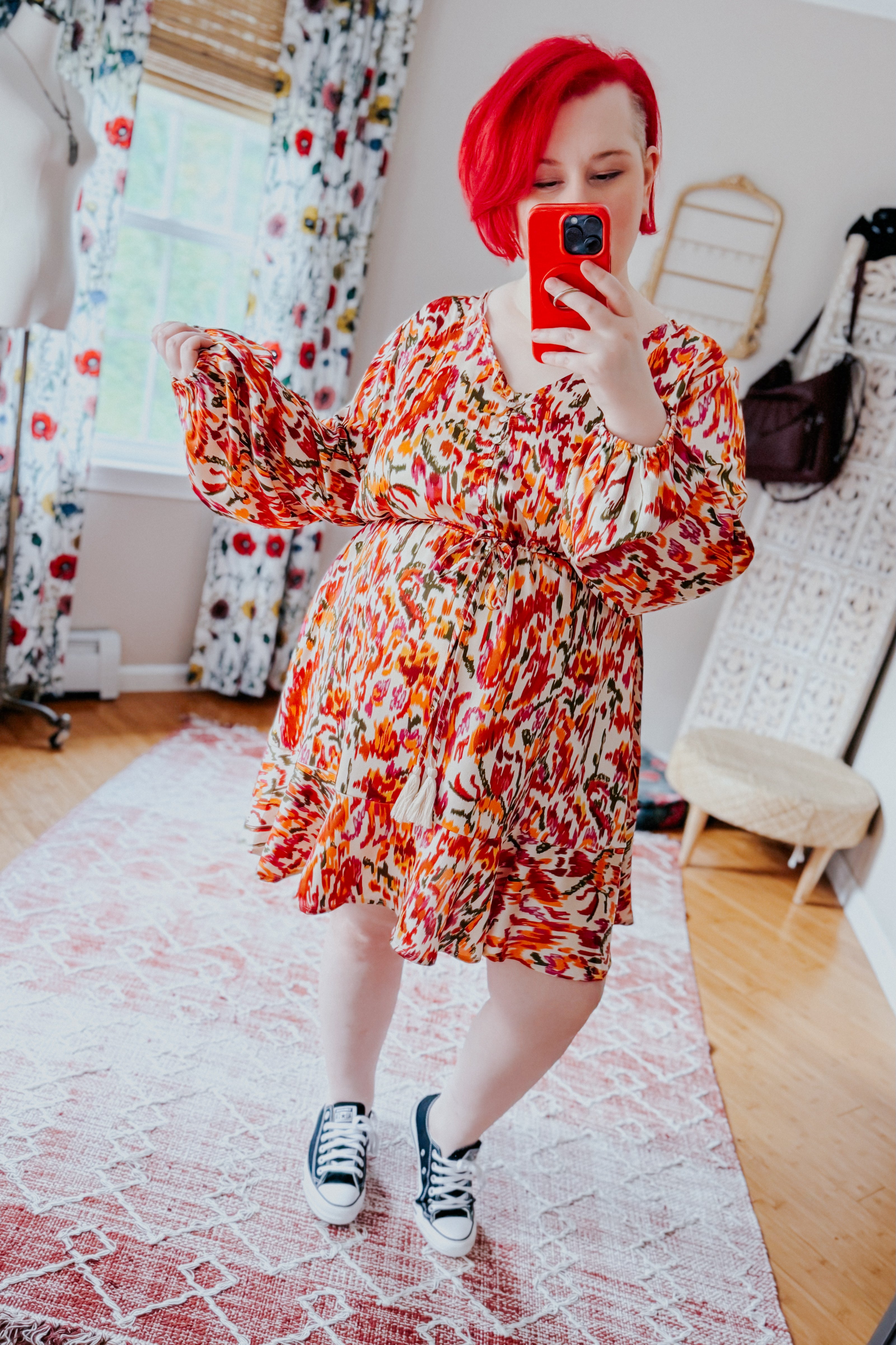 Woman with bright red hair wearing orange patterned V-neck dress with balloon sleeves and tie waist taking selfie indoors