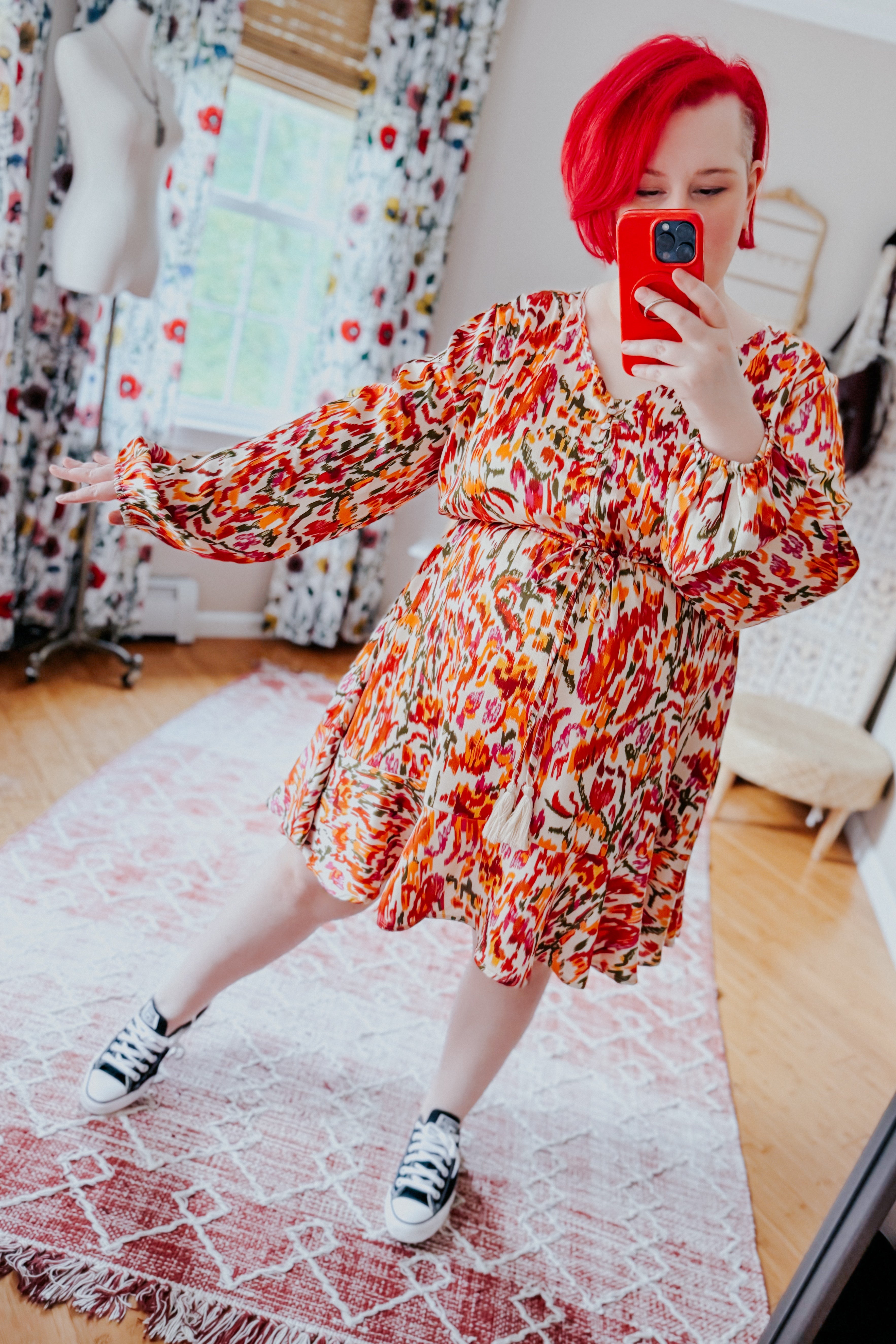 Woman with red hair wearing vibrant orange and red patterned V-neck balloon sleeve dress taking mirror selfie indoors
